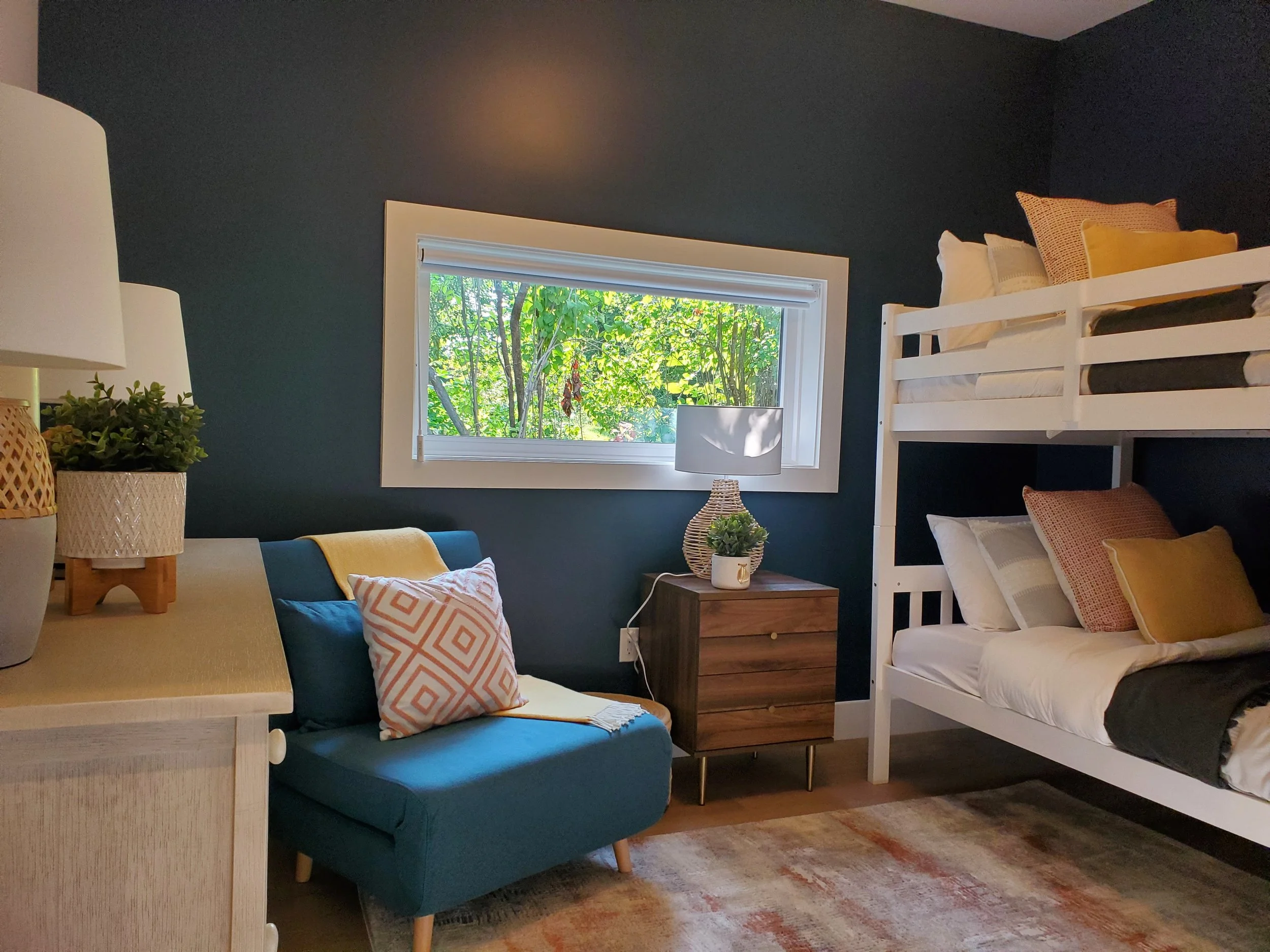 A cozy bedroom with dark blue walls, a window showing green foliage outside, a bedside lamp, a small wooden table with a potted plant, a dark green sofa with patterned pillows, and a white bunk bed with yellow and beige pillows.
