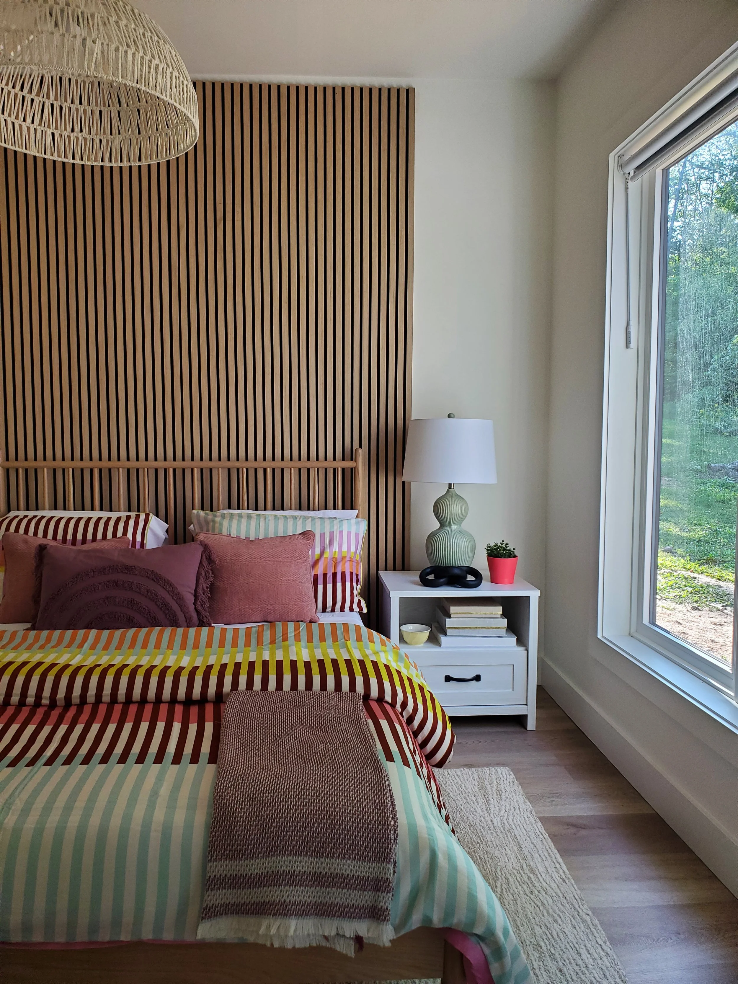 A bedroom with a bed covered in a striped comforter with various colors, and pillows in shades of purple and pink. A woven throw is placed at the foot of the bed. To the right, a white nightstand with a decorative lamp, a small plant in a red pot, an