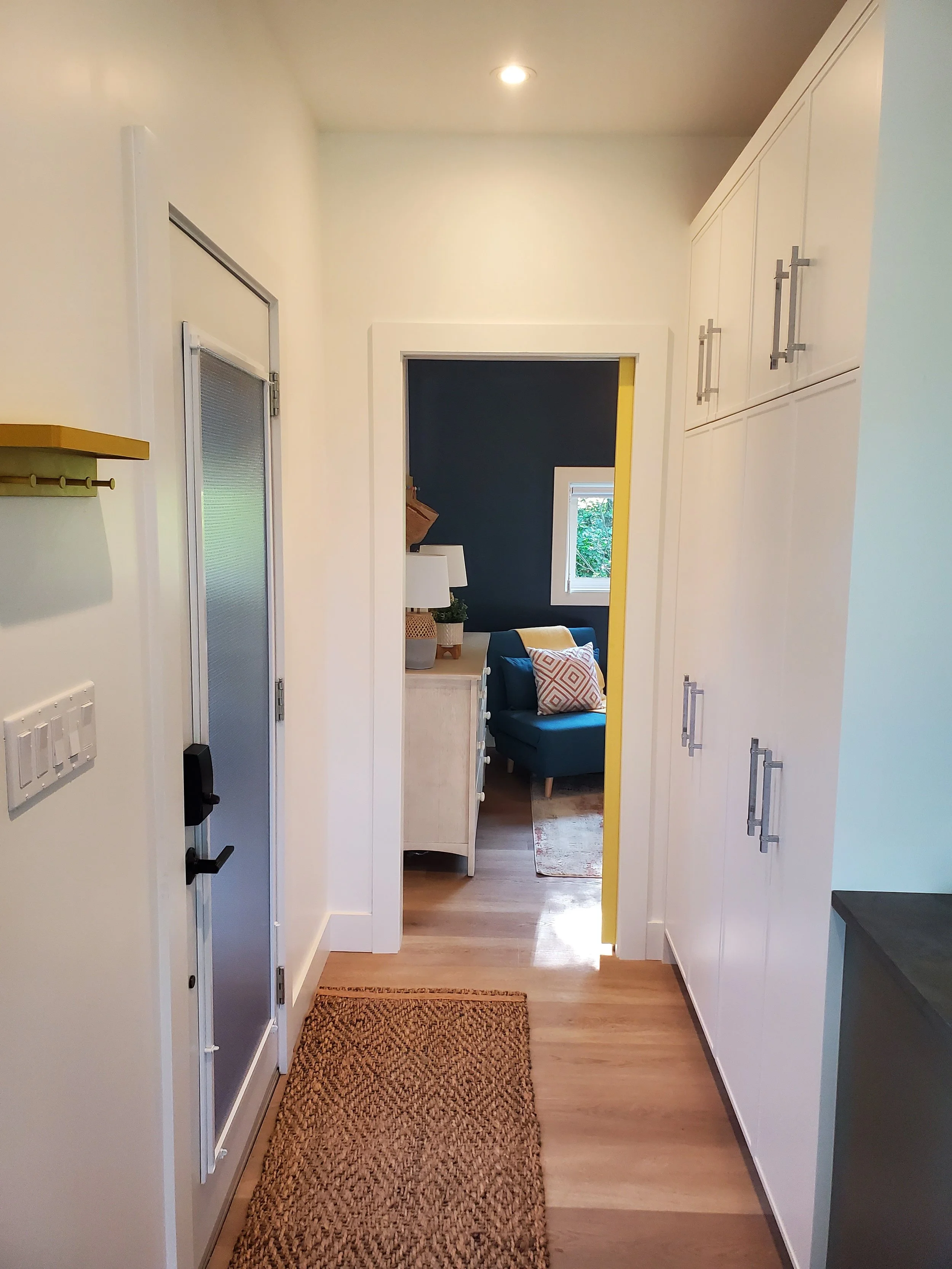 A hallway with white walls, a woven brown rug on a wooden floor, and white cabinetry on the right. At the end of the hallway, there's a room with dark blue walls, a window, and a seating area with a dark chair and decorative pillows. The doorway has 