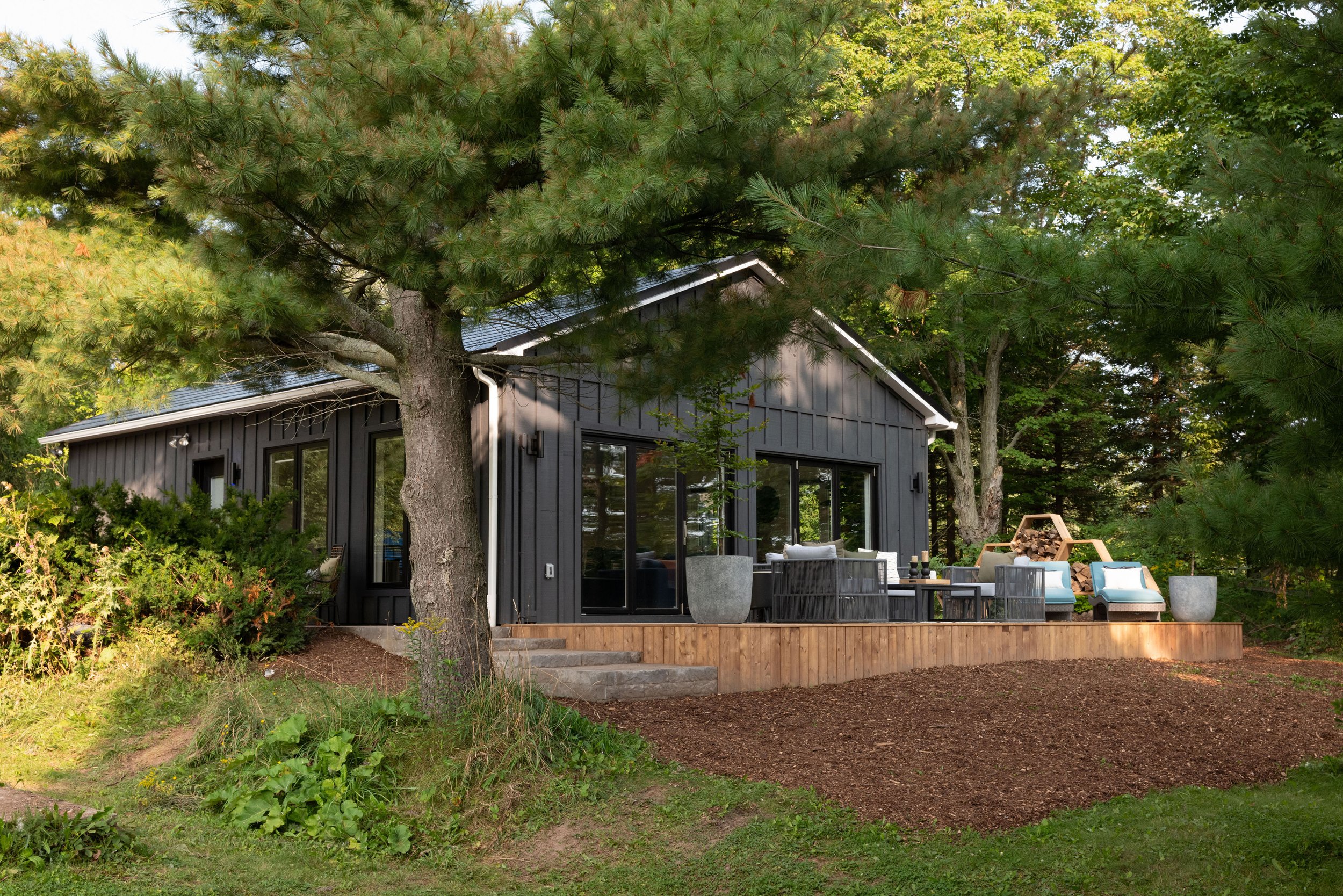 A modern black house with large glass windows is surrounded by tall trees, including pine trees, on a green lawn. There is outdoor patio furniture with chairs and a table, and a firewood storage area in the backyard.