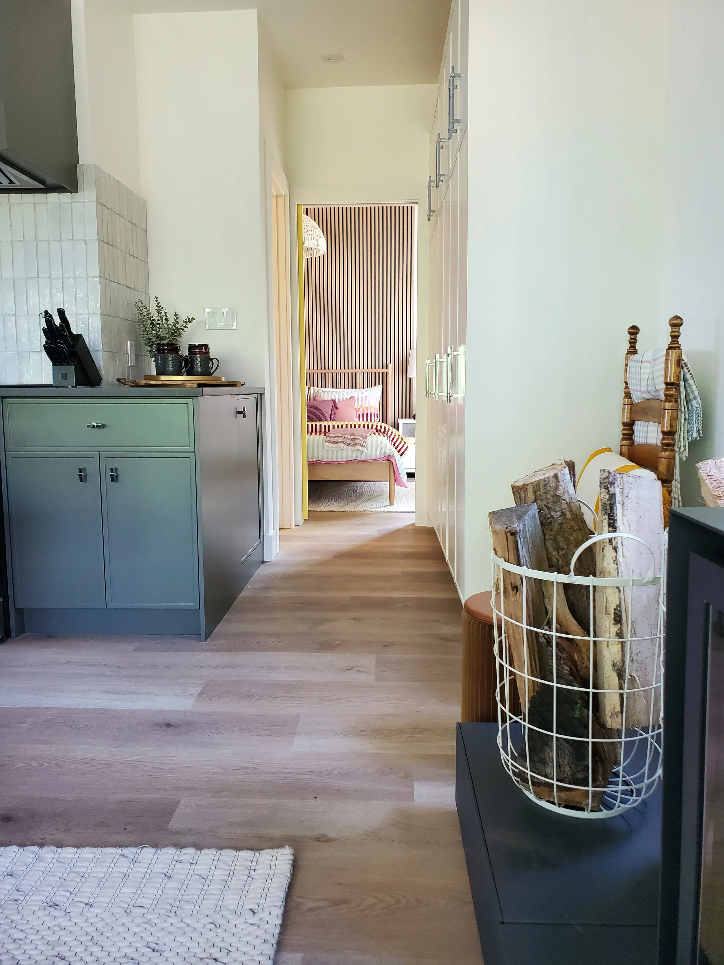 Interior view of a cozy living space with a doorway leading to a bedroom in the background. The room features a light-colored wood floor, a green cabinet on the left with kitchen utensils and decorative jars, a black table or stand with firewood in a