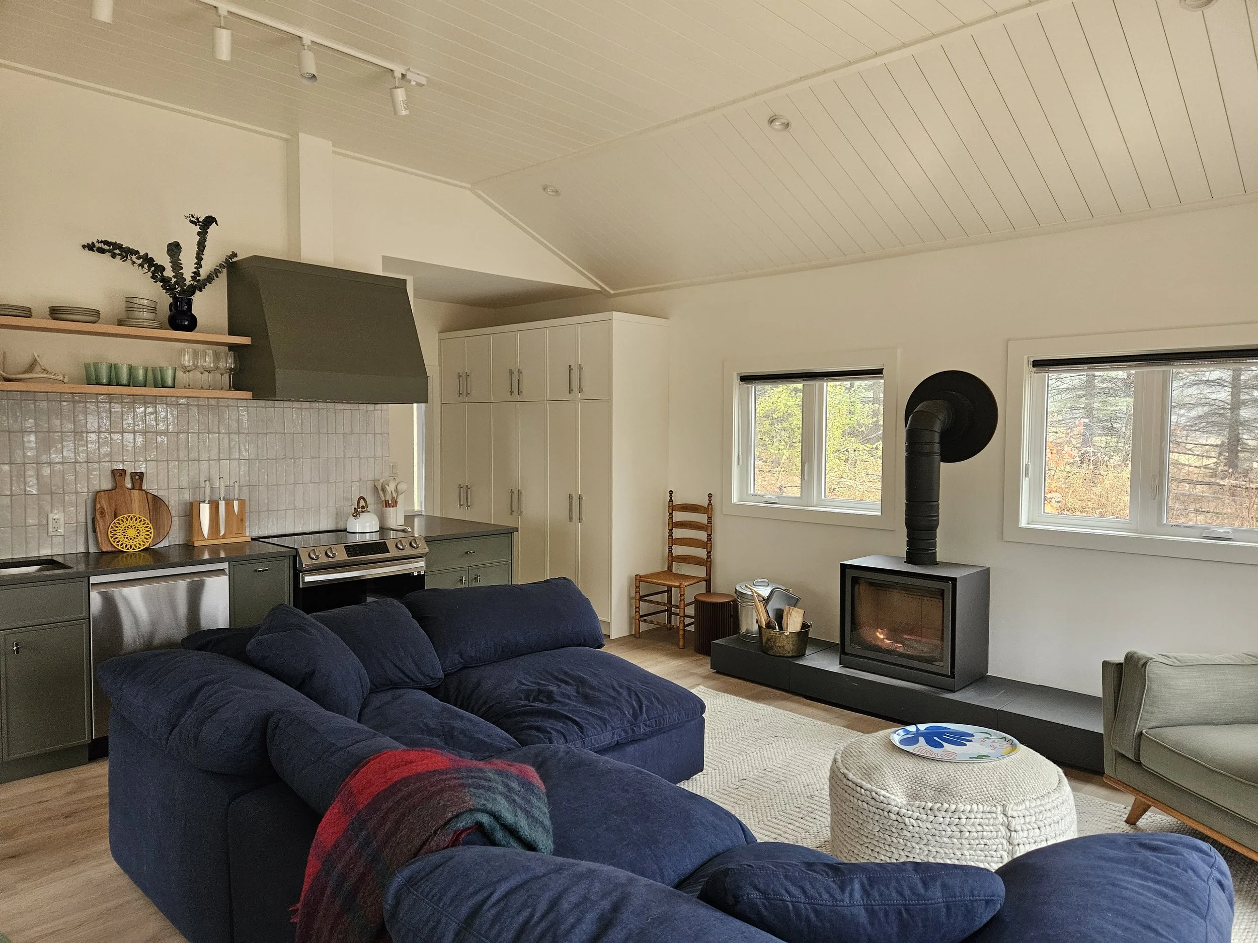 A cozy living room with a blue sofa, a white ottoman, a fireplace, and several windows showing trees outside.