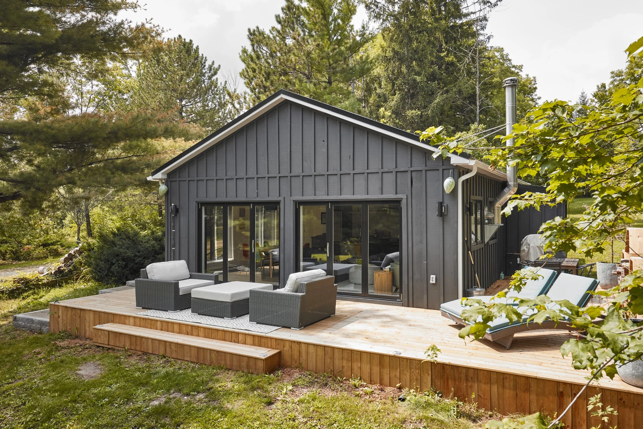 A modern house with large glass sliding doors opening onto a wooden deck with outdoor furniture, surrounded by trees and greenery.