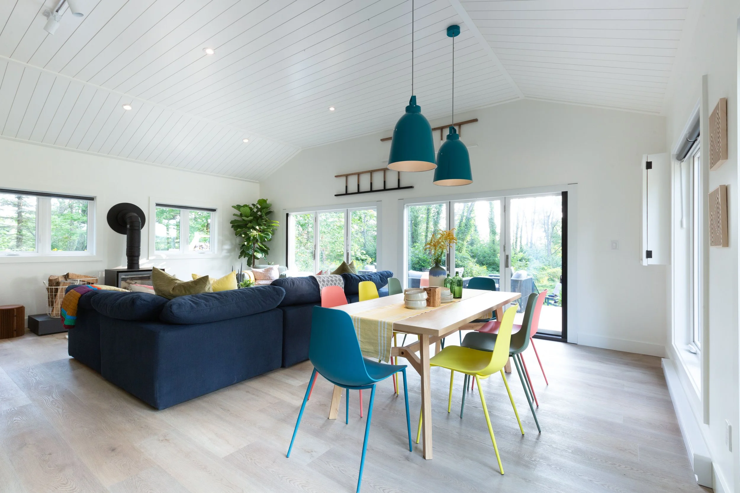 Bright living and dining room with white vaulted ceiling, blue pendant lights, large windows, hardwood floors, navy sofa, colorful chairs, and a wooden dining table with a yellow table runner.