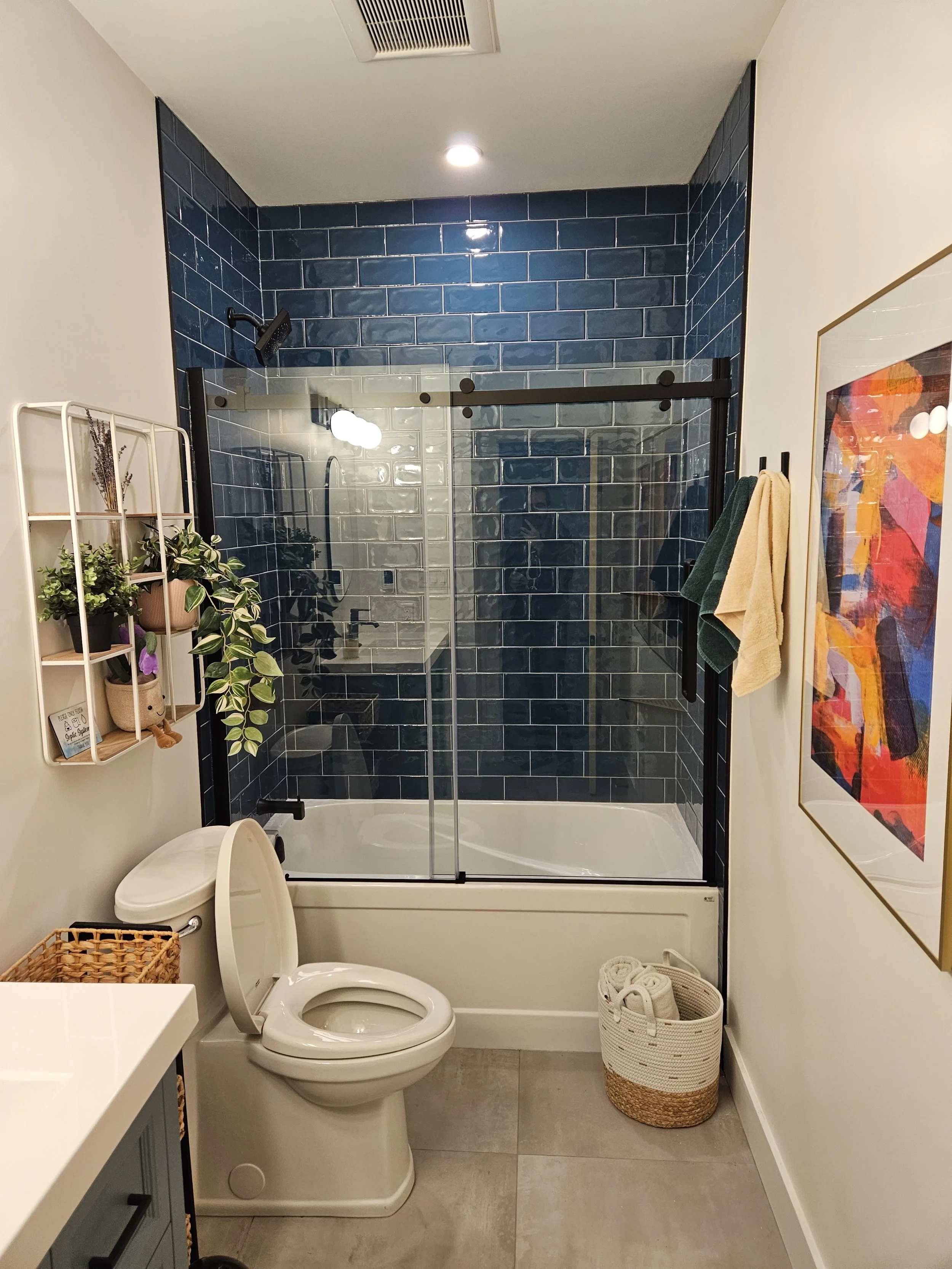 A modern bathroom with a walk-in shower with blue subway tiles, a glass door, a toilet, a white counter with cabinet drawers, a small white shelf with plants and decor, a framed abstract colorful art on the wall, and various towels and baskets for or