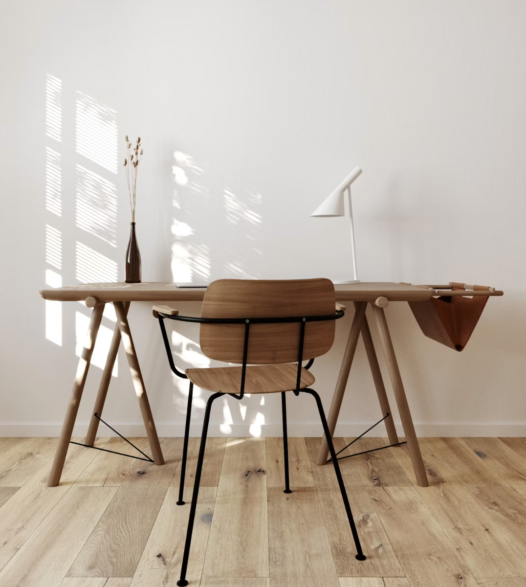 Minimalist desk setup with wooden table, black metal-framed wooden chair, white desk lamp, and a tall vase with dried plants, against a white wall with sunlight and shadow patterns.