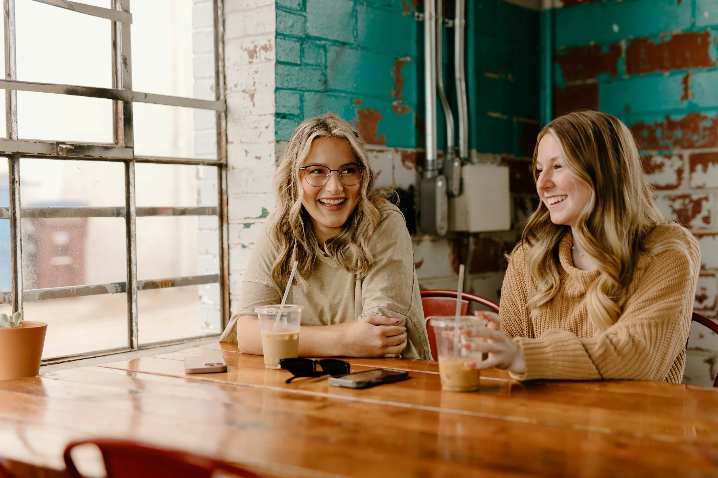 Two women with long, wavy hair and glasses sitting at a wooden table in a cafe, smiling and talking to each other.
