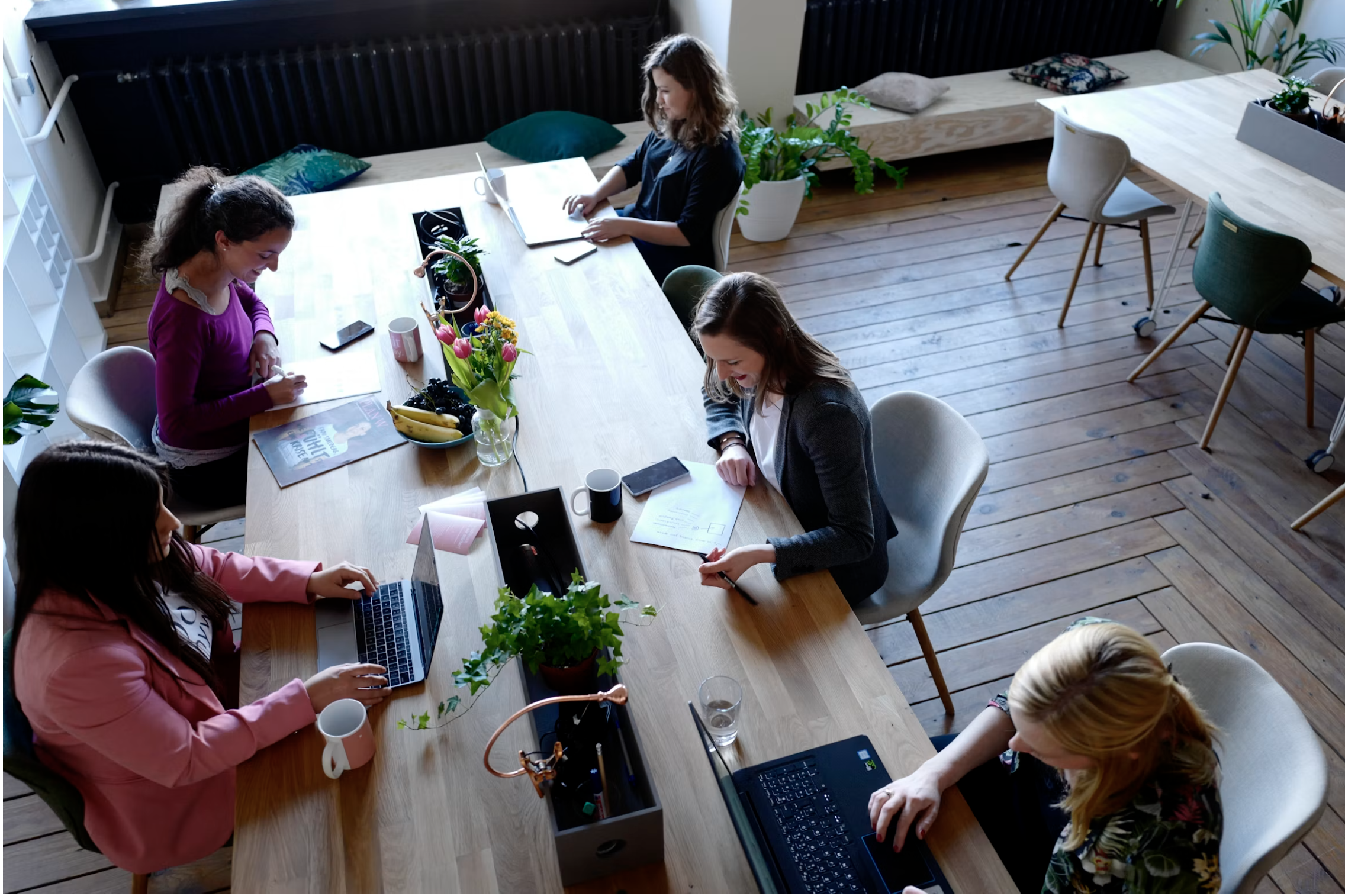 A group of women sitting around a wooden table in a bright, modern room with wooden floors, working on laptops and taking notes, with flowers and plants on the table.