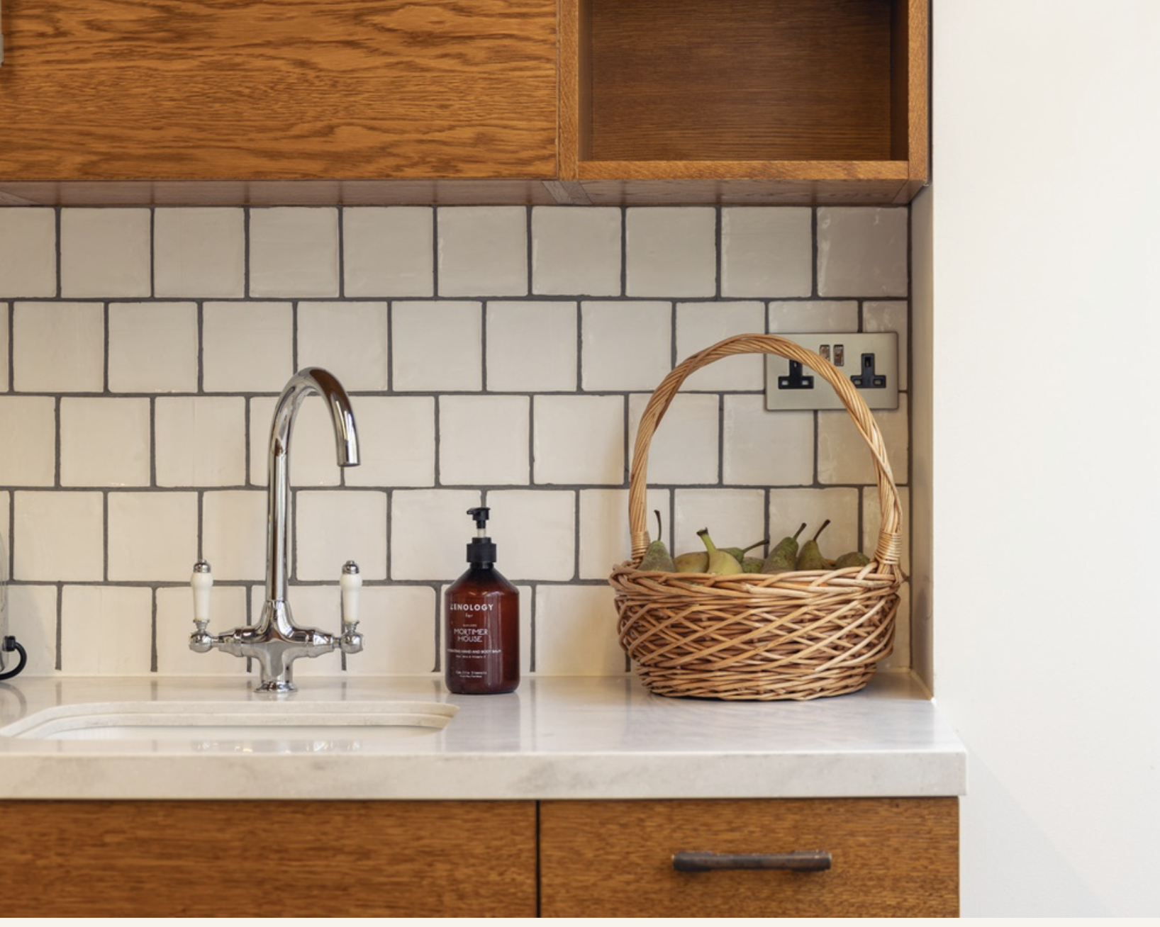 Kitchen countertop with a stainless steel faucet, a brown soap dispenser, a wicker basket with pears, and a white tiled wall with electrical outlets.