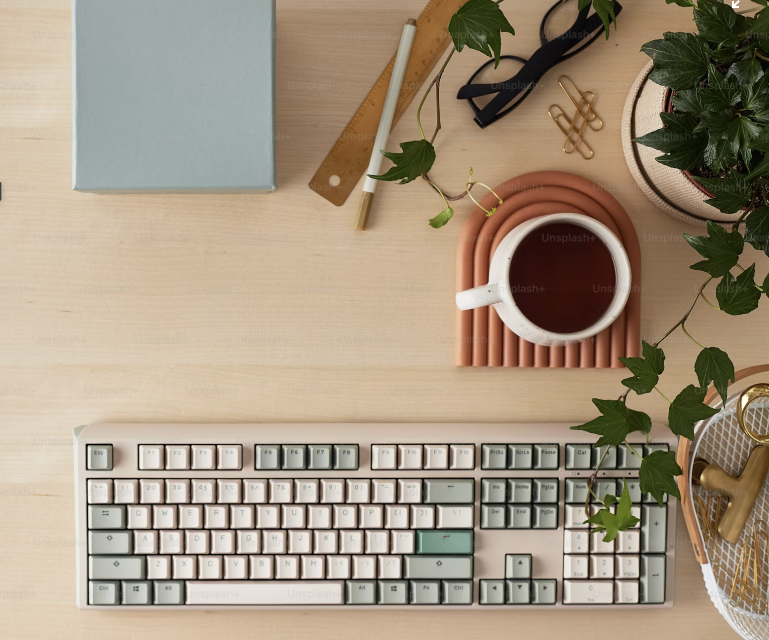A workspace with a computer keyboard, a cup of tea on a pink tray, a potted plant, a notebook, a pen, a ruler, glasses, and gold pushpins on a light wooden desk.
