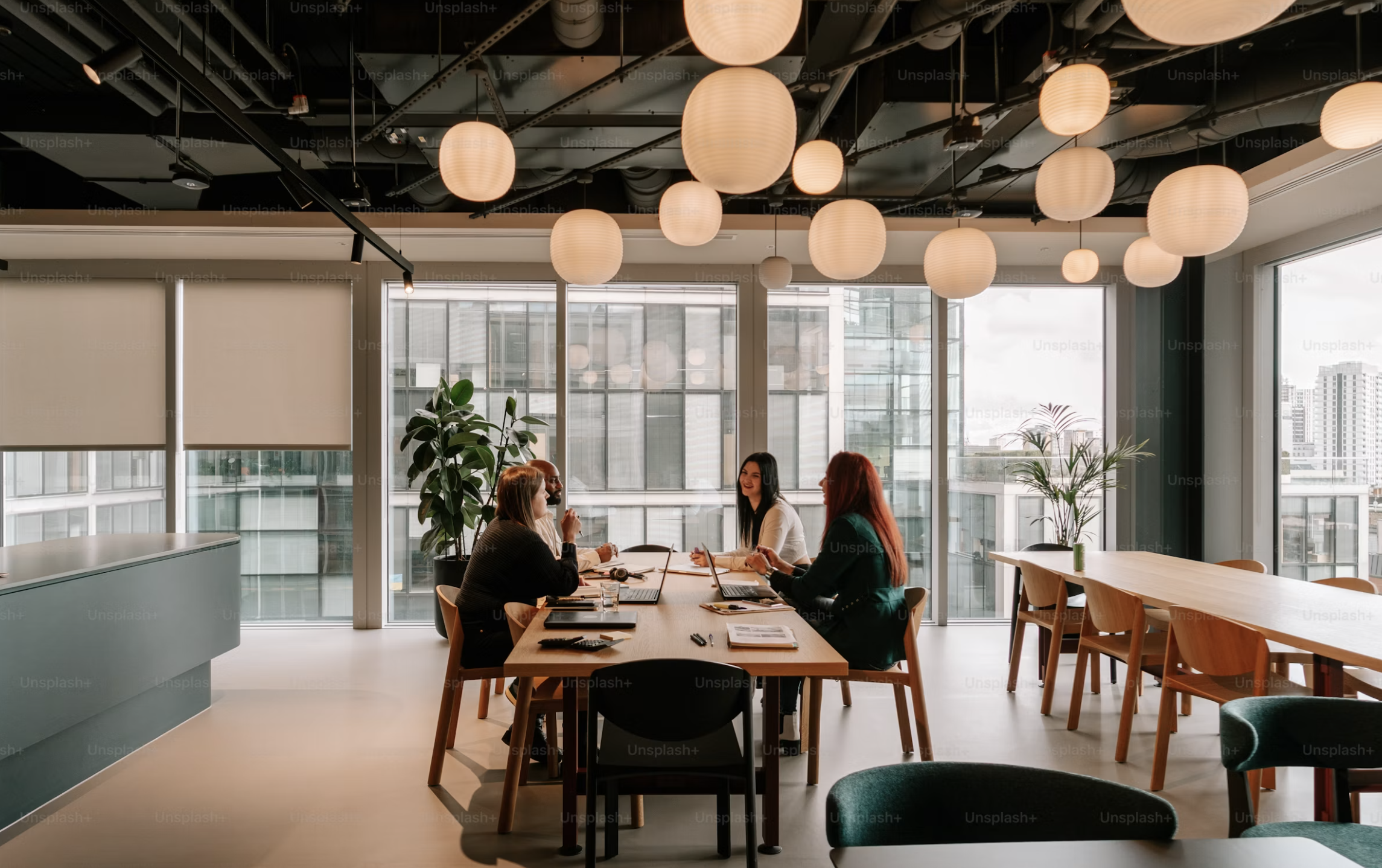 Four people sit at a table in a bright, modern office space, engaged in conversation, with laptops, notebooks, and pens on the table. The room has large windows with views of city buildings, potted plants, and paper lantern ceiling lights.