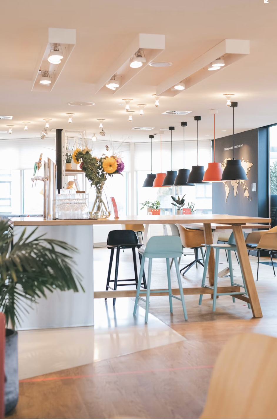 Modern cafe interior with a tall wooden table, colorful chairs, pendant lights, and a flower arrangement on the table.