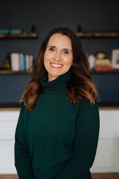 Woman with shoulder-length brown hair smiling in a dark green turtleneck sweater, standing in a room with a bookshelf in the background.