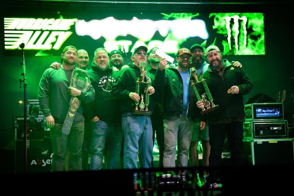 Group of eight men on stage holding trophies and a championship belt, celebrating in front of a green illuminated "Turtle Alley" sign, with audio equipment visible.