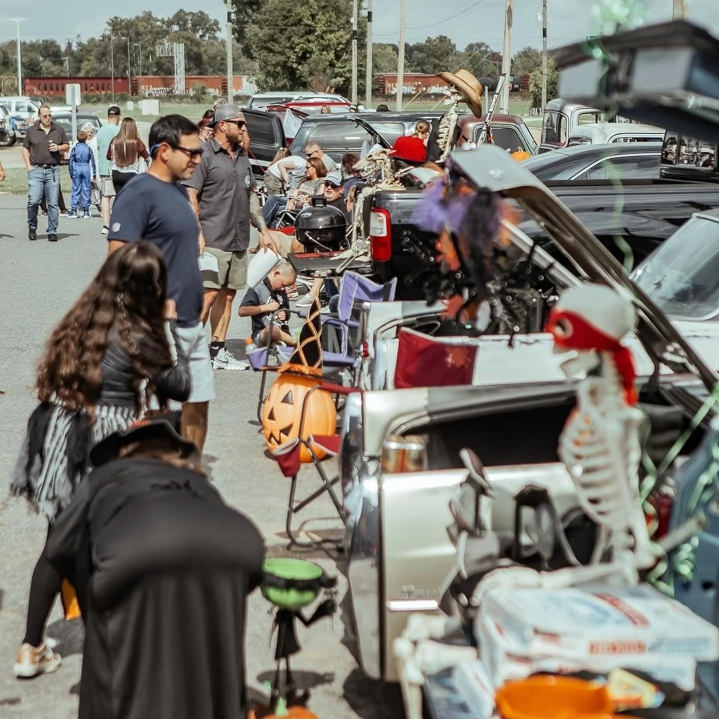 People browsing Halloween decorations and costumes at an outdoor market, with decorated items such as a pumpkin bucket and a skeleton in the foreground.