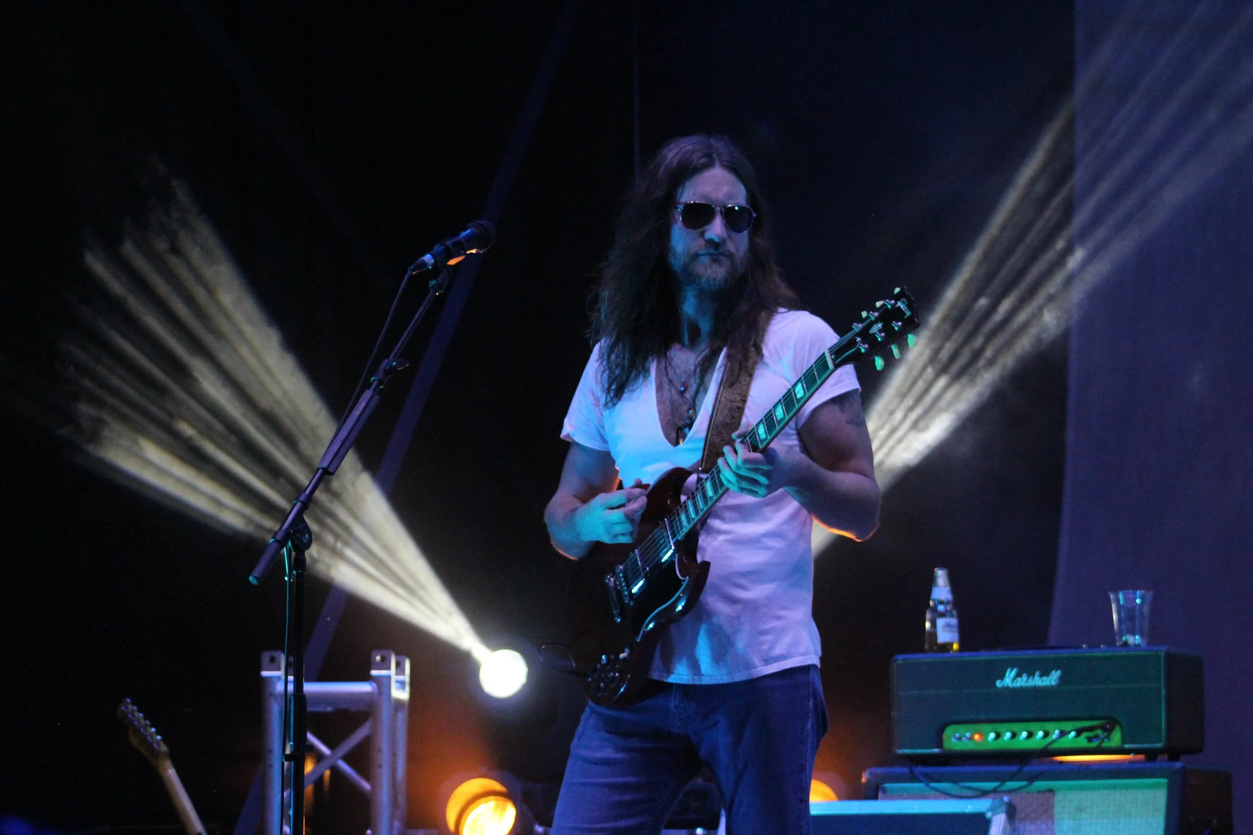 A long-haired man wearing sunglasses and a white T-shirt playing an electric guitar on stage with stage lighting behind him.
