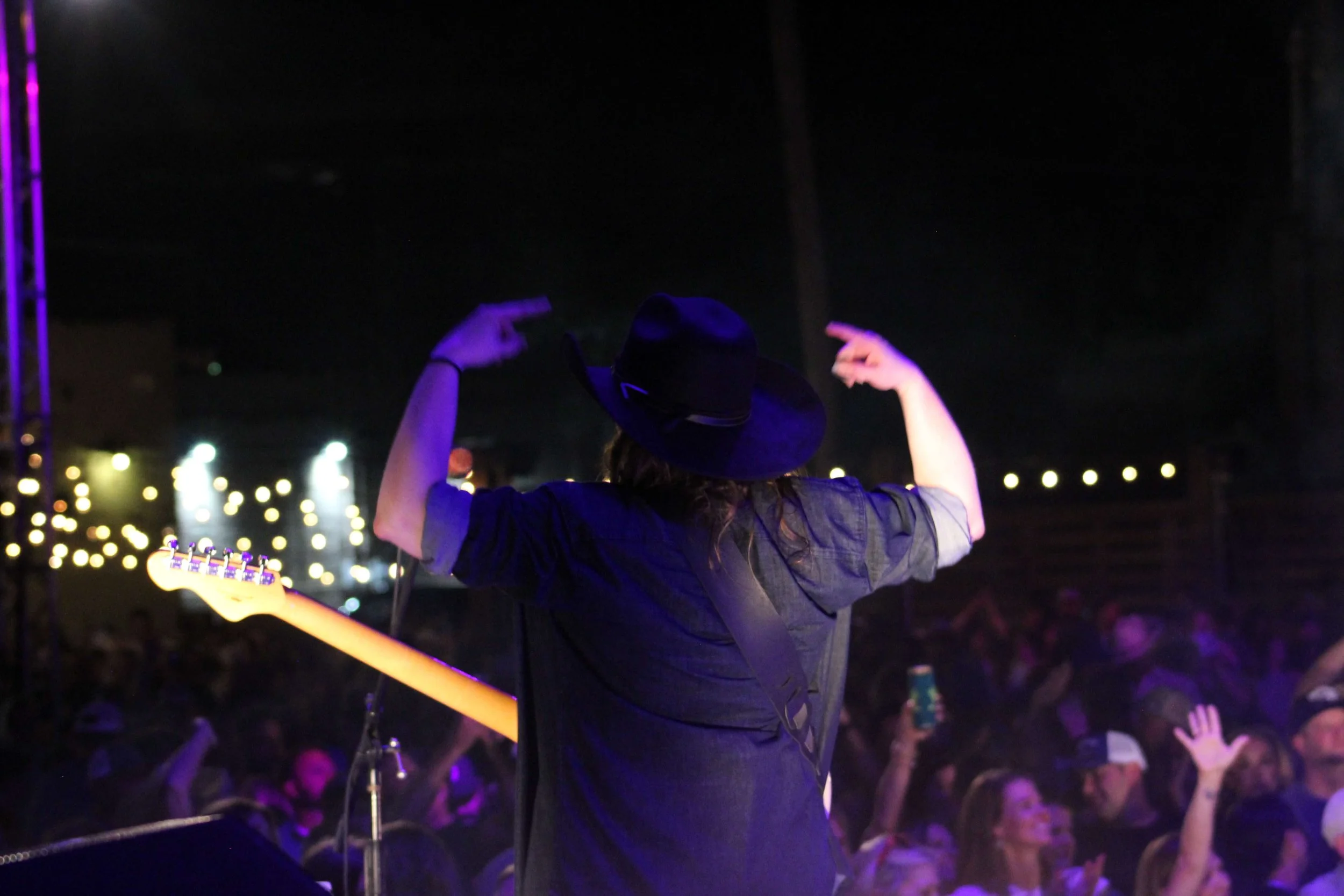 A musician wearing a hat is performing on stage at night, holding a guitar, with an audience in front and string lights in the background.