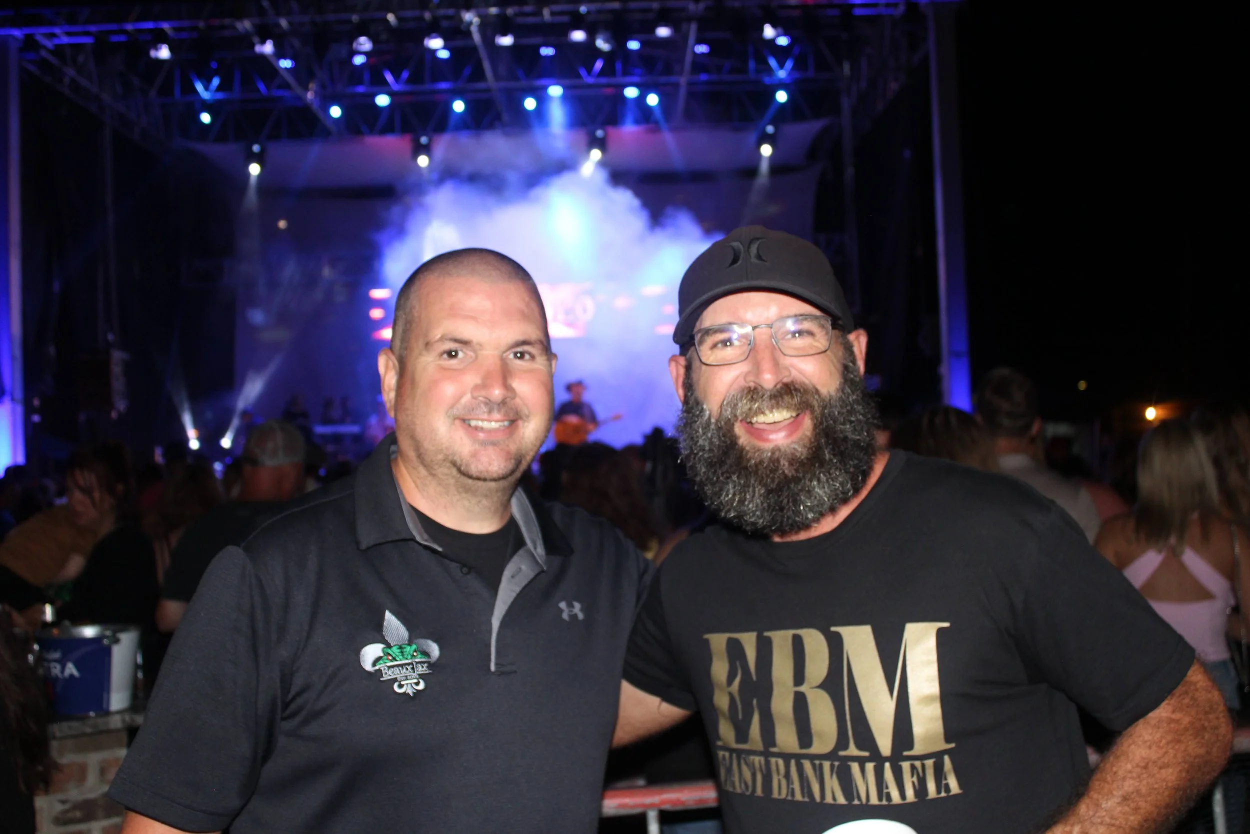 Two men smiling at a concert or event with a stage and blue lights in the background. One is wearing a black polo shirt with a logo, and the other is wearing glasses, a black cap, and a black T-shirt with graphic text.