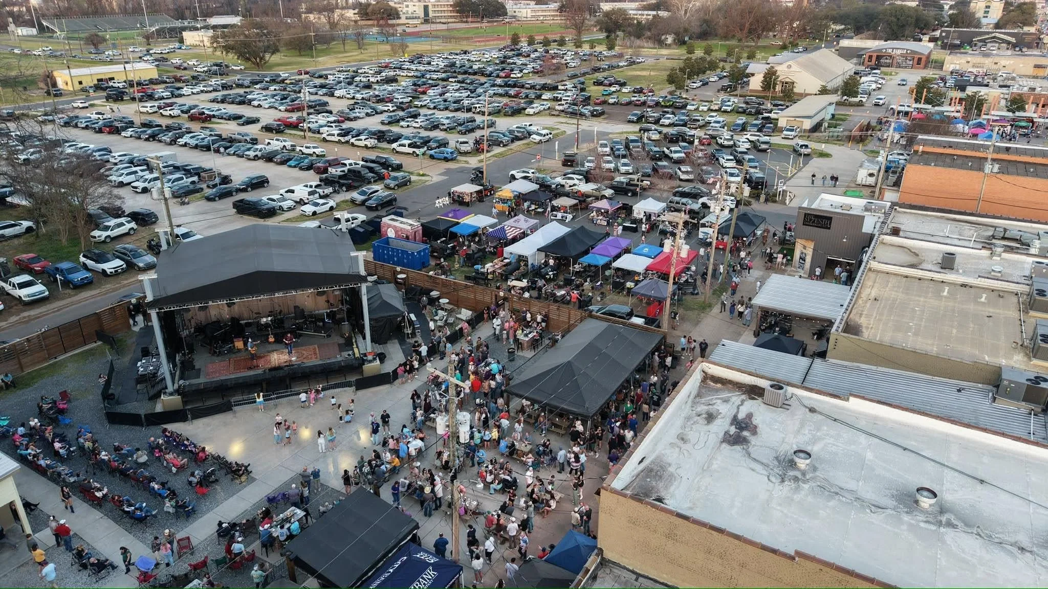 An aerial view of a crowded outdoor event with a stage and tents, surrounded by a parking lot filled with cars.