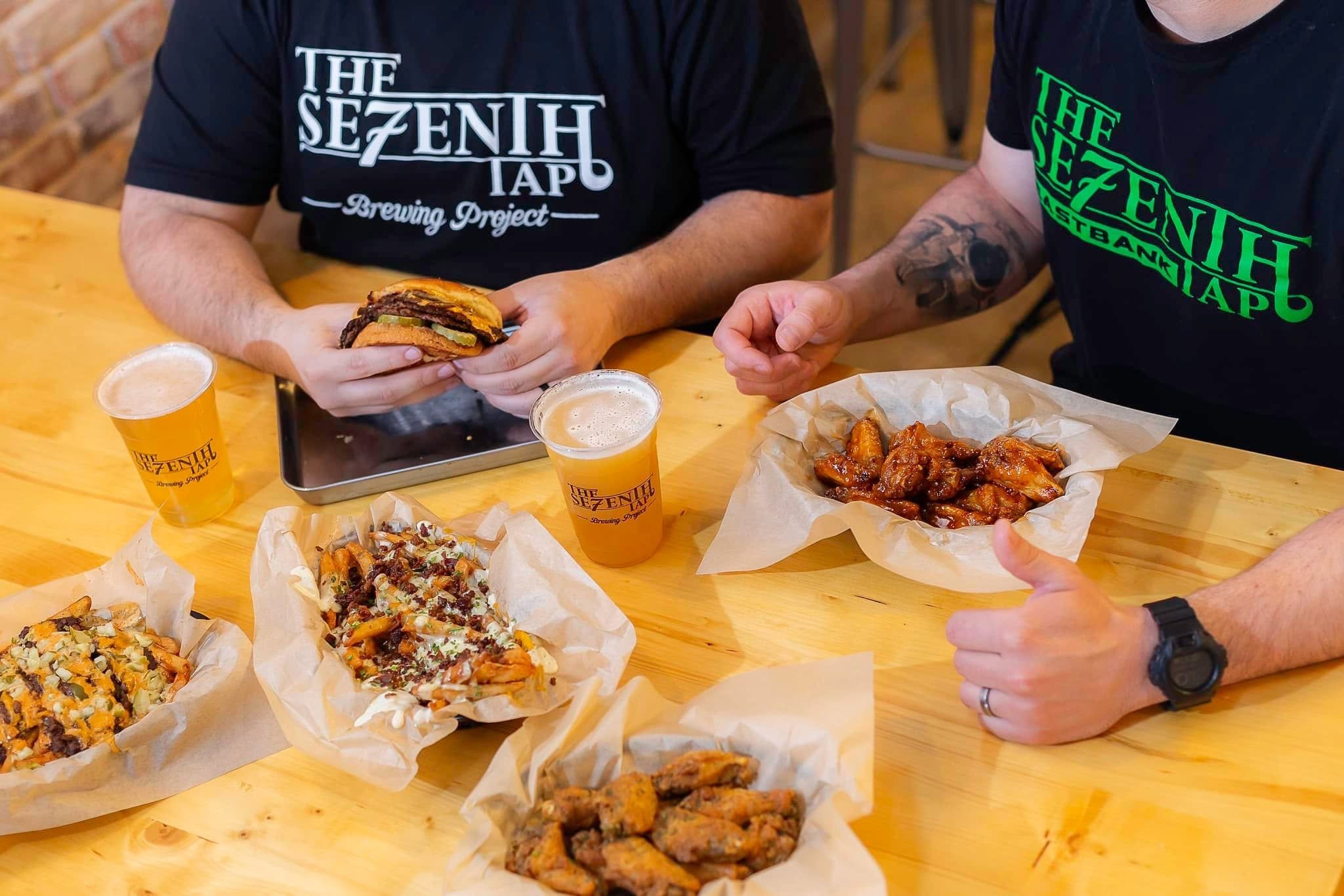 Two people sitting at a wooden table holding food, with pizza, fried chicken, and drinks in front of them. They are wearing black T-shirts with green and white text that reads 'The Sezenh Tap Brewing Project' and 'The Sezenh Tap'.