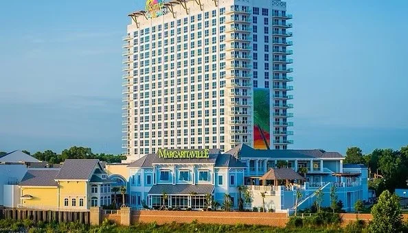 A tall hotel building with a colorful sculpture on top, and a multi-colored mural on one side. Smaller blue and yellow buildings in the foreground with a sign that reads 'Margaritaville'.