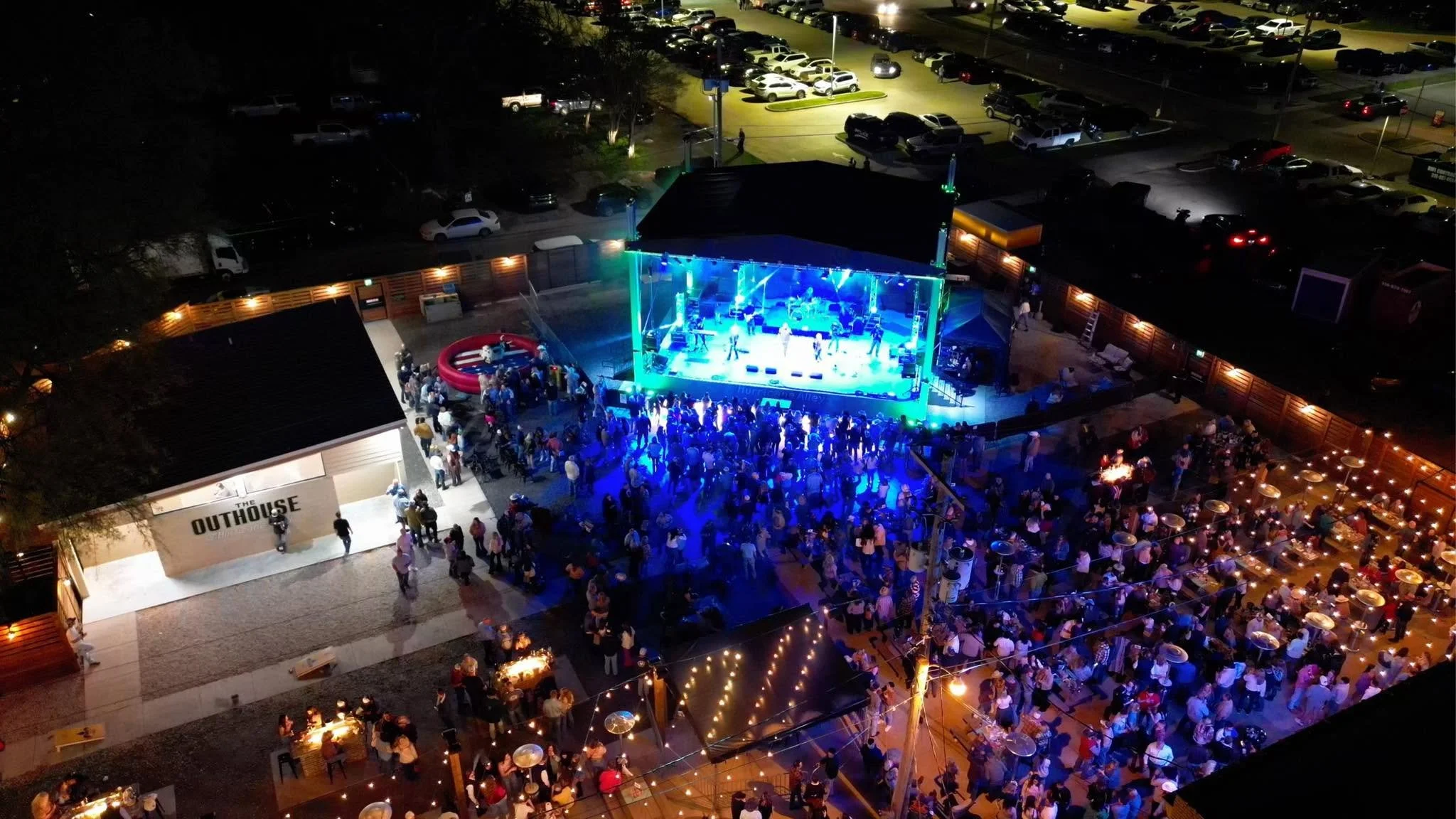 Nighttime outdoor concert at a venue called 'The Outpost.' A large stage with blue lighting hosts a live band, and a crowd gathered in front. Surrounding the stage are various seating and standing areas with people enjoying the event, some near a small inflatable pool. Parking lots and illuminated walkways are visible in the background.