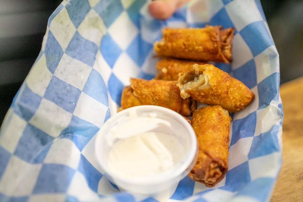 Four fried egg rolls with a side of white dipping sauce in a paper basket lined with blue and white checkered paper.