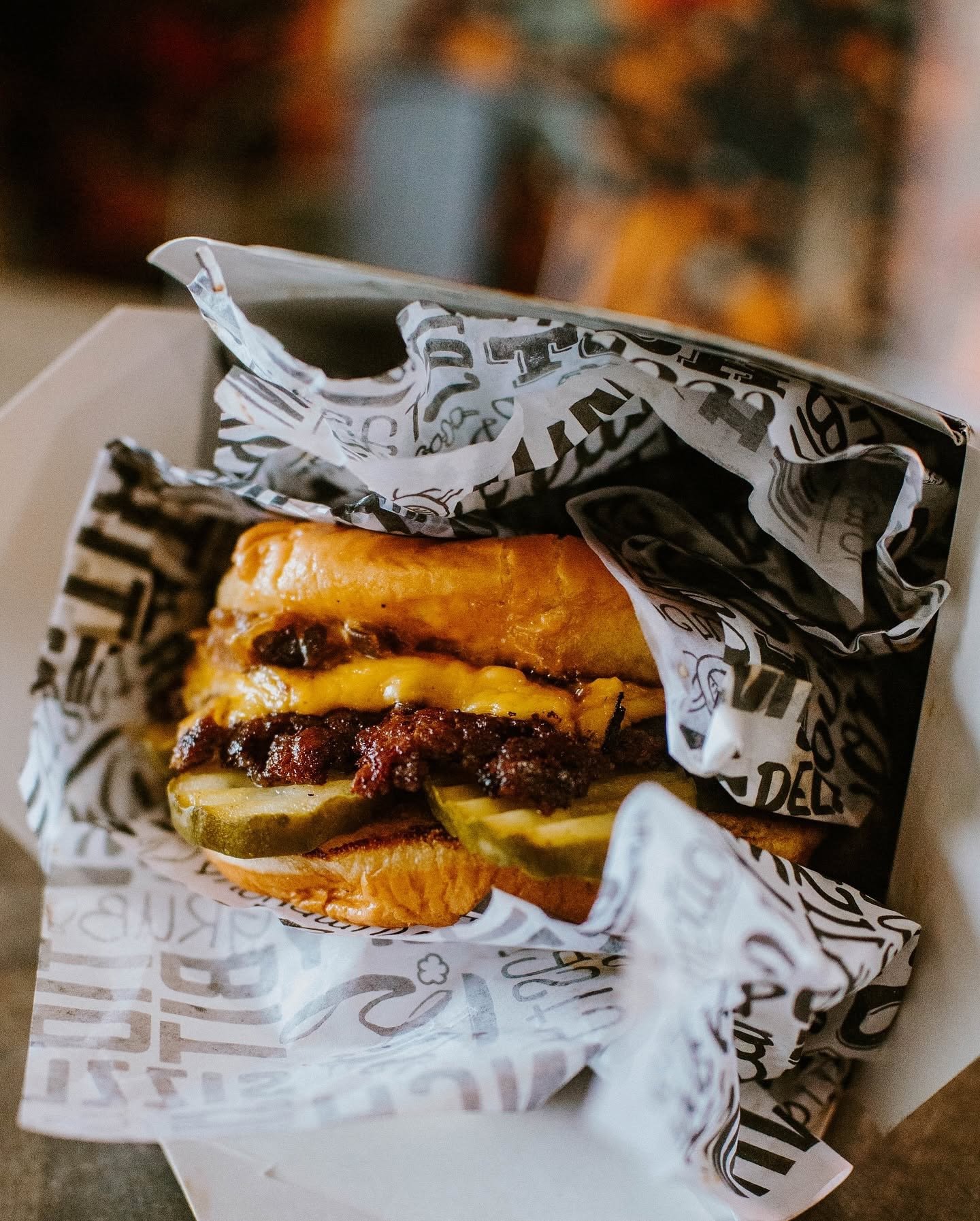 A cheeseburger with pickles and bacon inside a paper wrapper, partially visible, in a white box.