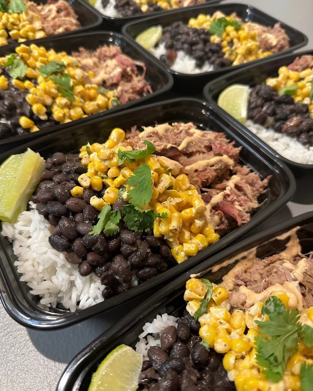 Multiple meal prep containers filled with white rice, black beans, shredded meat topped with corn, garnished with cilantro, and lime wedges.