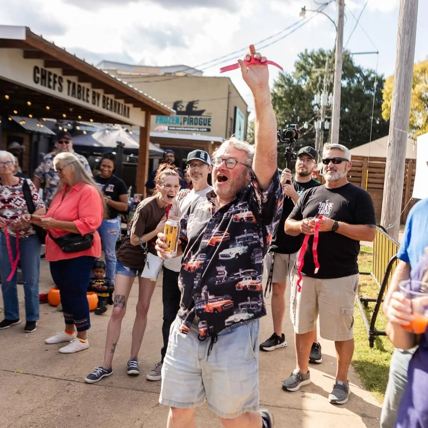 A man in a car-themed shirt holding a ribbon with a crowd of people around him. The crowd is laughing and smiling. Some are holding ribbons, and a woman is looking at her phone. There is a sign that reads 'Chefs Table by Bellavia' and a storefront in the background.