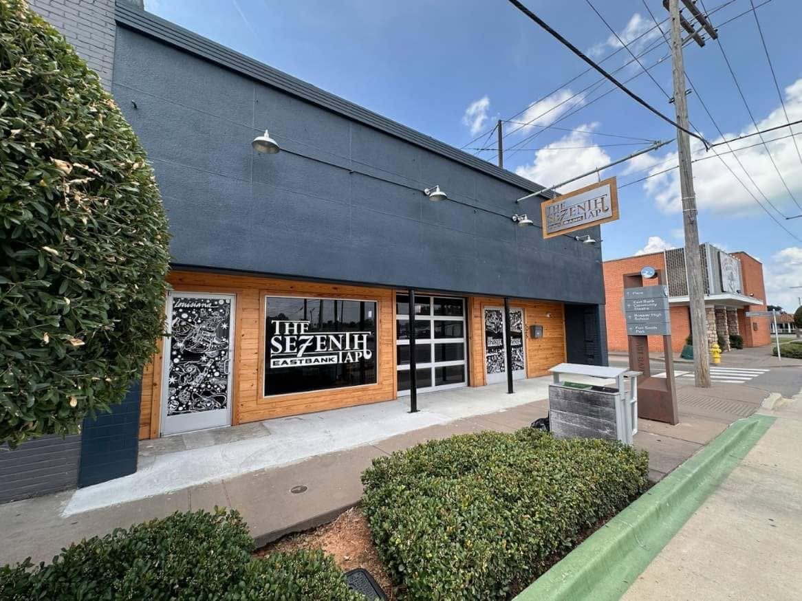 Exterior view of The Zen House restaurant with black upper walls, wooden accents, a large window, and signage on the building and an overhead sign, with a sidewalk, bushes, and utility poles in the scene.