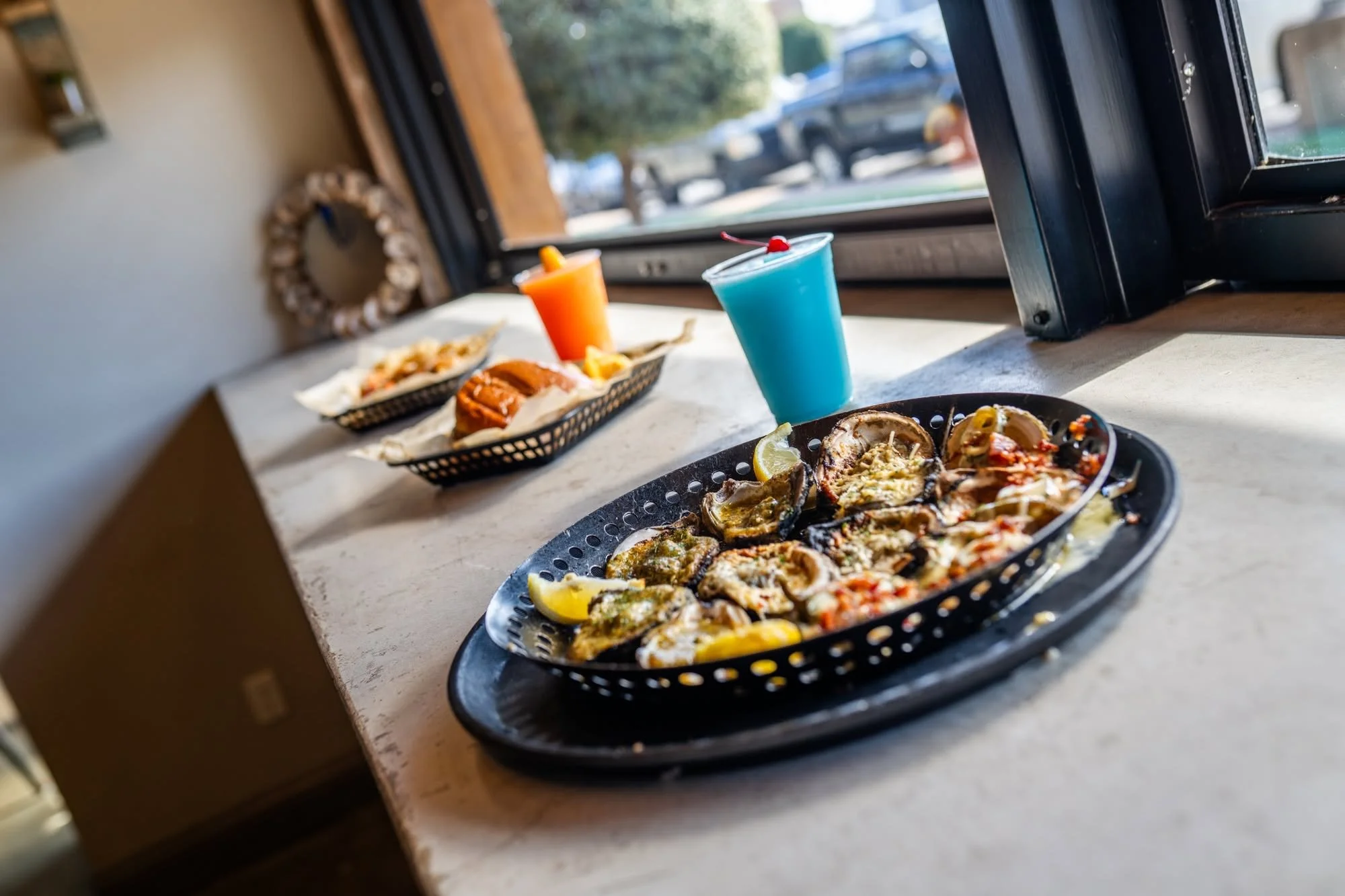 A black plastic tray of baked oysters with lemon wedges and seasoning on a white countertop, with a plate of tacos and colorful drinks in the background near a window.