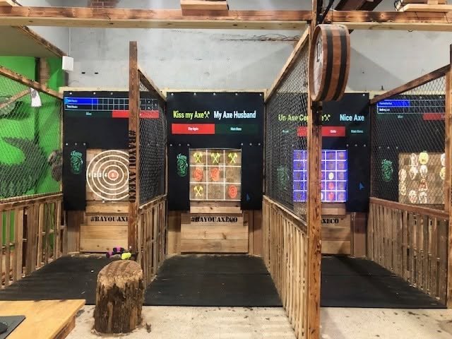 Indoor axe-throwing range with three lanes, each equipped with digital scoring displays and targets. The lanes are separated by wooden and wire fencing, with a log used as seating in the foreground.