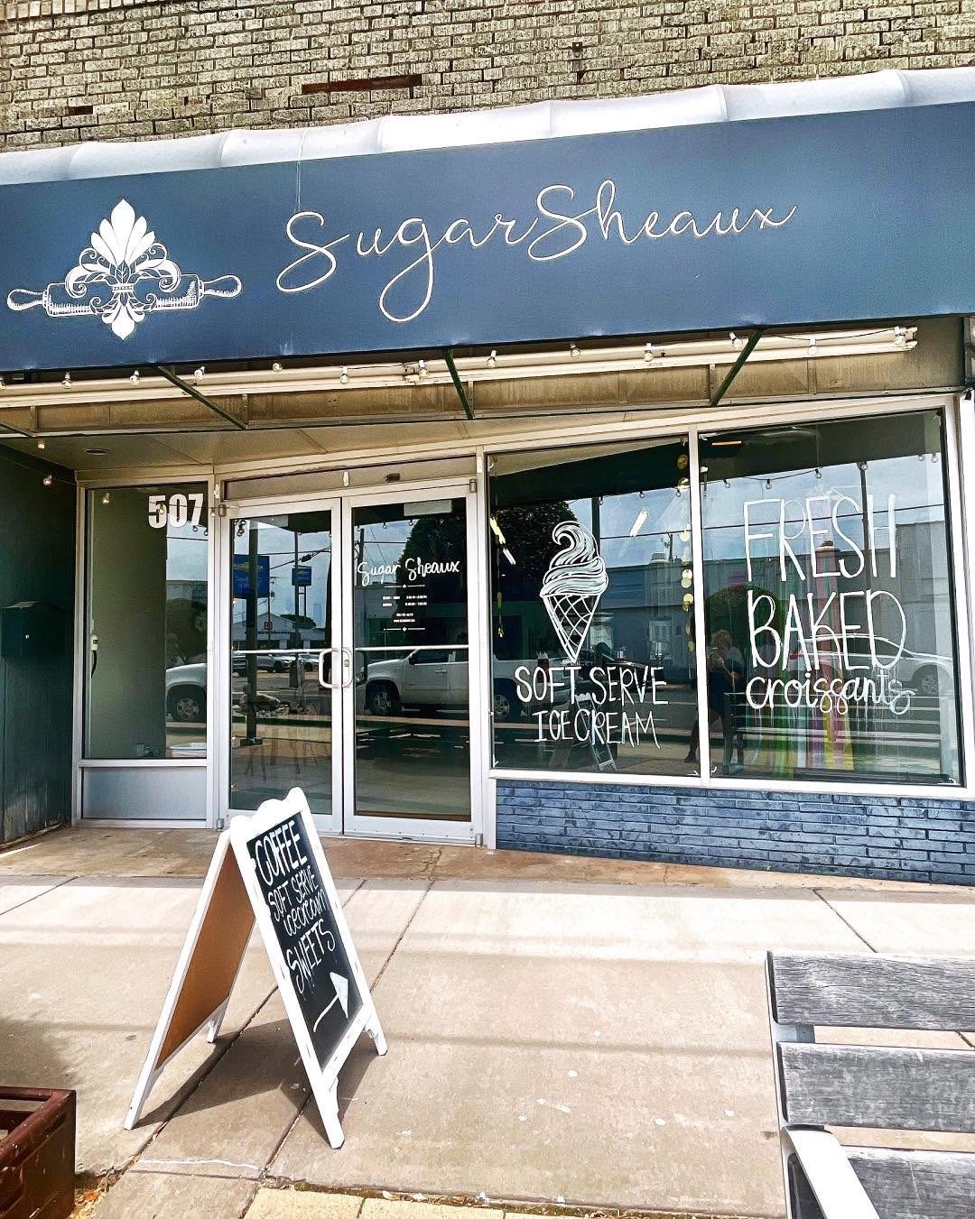 Front view of Sugar Sheaux bakery storefront with a blue awning, large glass windows, and white handwritten signs advertising ice cream, croissants, and baked goods. There is a black sandwich board on the sidewalk promoting coffee, sweets, and ice cream. A bench is partially visible on the right.