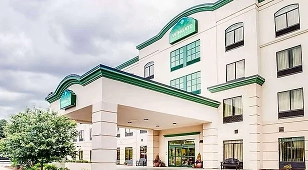 Exterior of a modern hotel named Wingate with white facade and green accents, featuring an entrance with a covered drop-off area and several windows, surrounded by greenery.