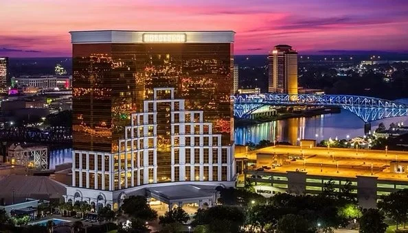 Nighttime city skyline featuring a large reflective hotel building, a bridge over a river, and illuminated city lights with a colorful sunset sky.