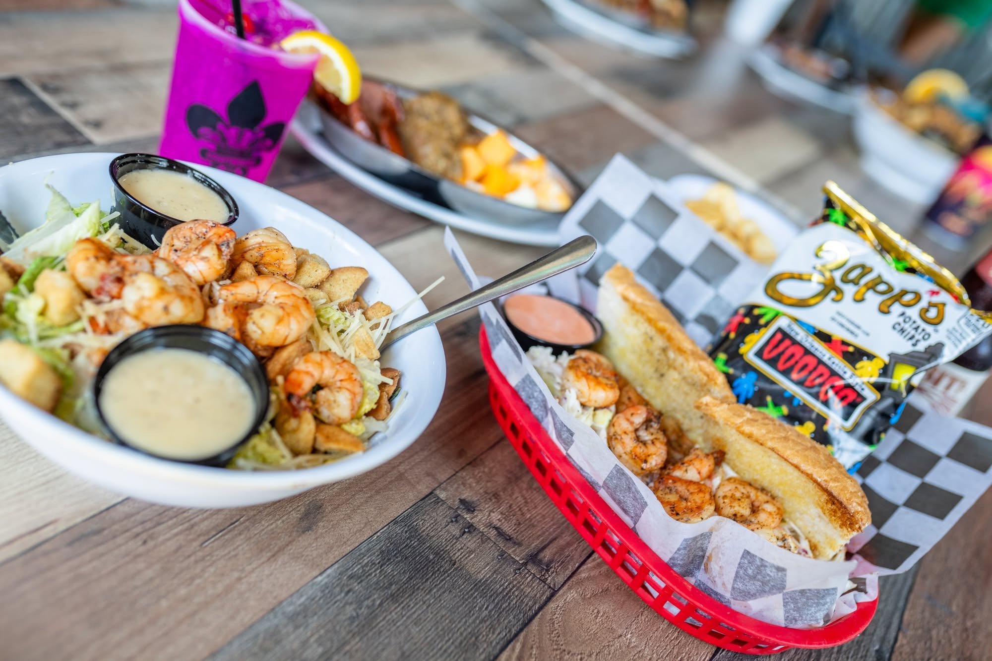 A tray of fried shrimp and a baked potato with toppings on a checkered paper, with a pink cup and a bowl of dipping sauce, on a wooden table.
