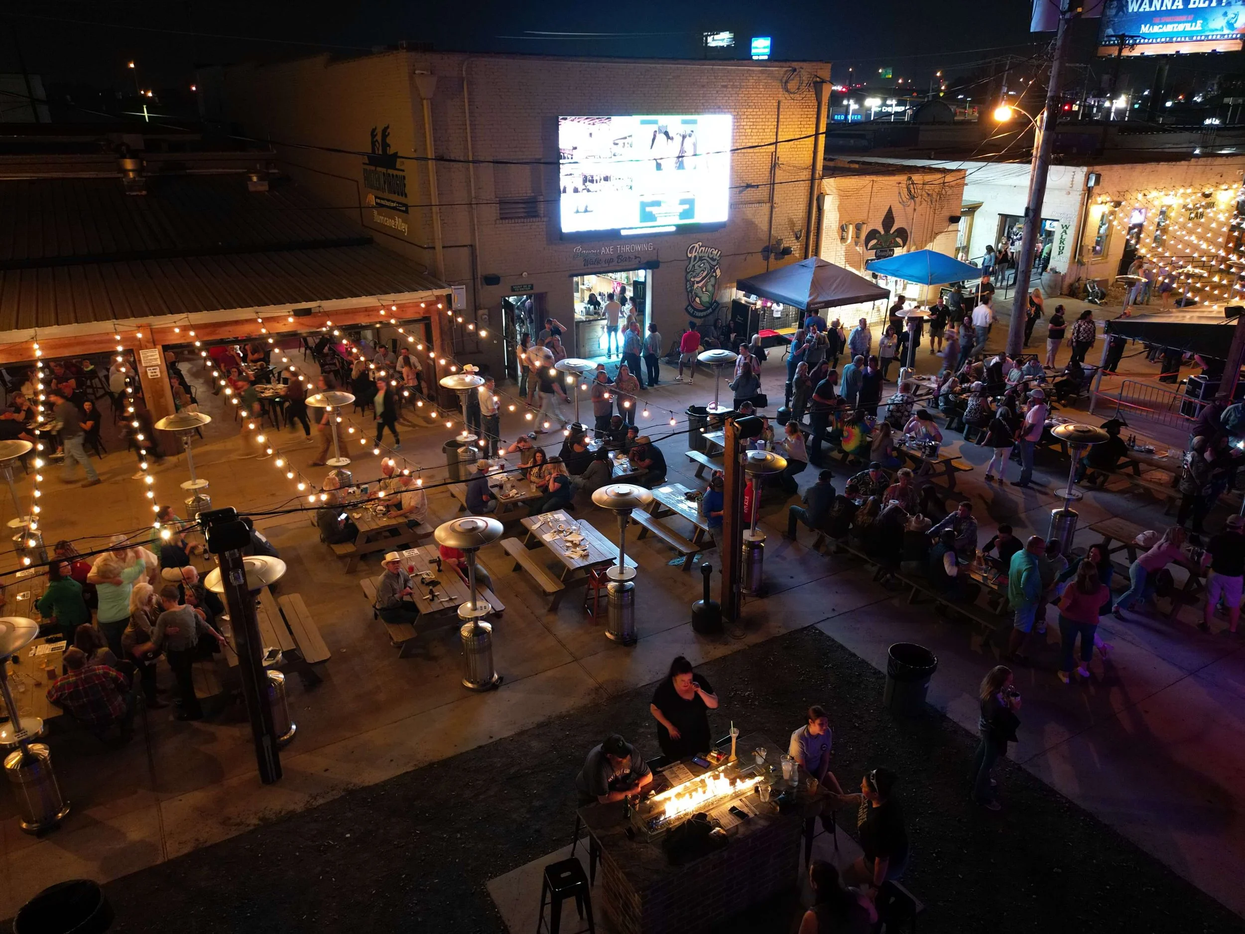 Night scene of an outdoor bar with string lights, people socializing and dining, brick buildings, a large illuminated digital billboard, and outdoor heaters.