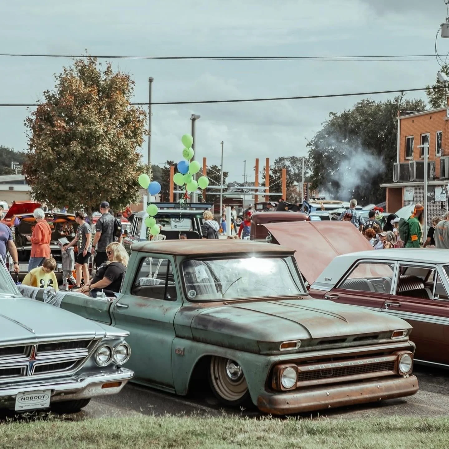 A vintage car show with old cars parked and people walking around, some looking at the cars, with a tree, balloons, and buildings in the background.