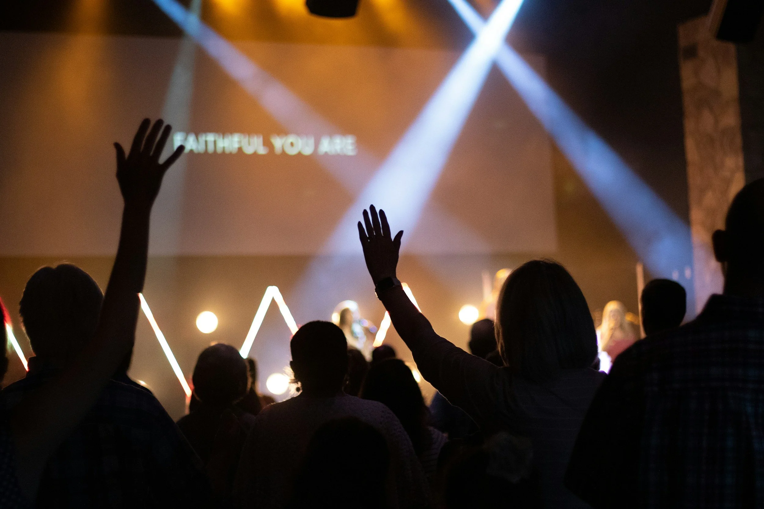 A congregation of people with raised hands attending a worship service or concert, with stage lights and a projection screen displaying the words 'FAITHFUL YOU ARE' in the background.