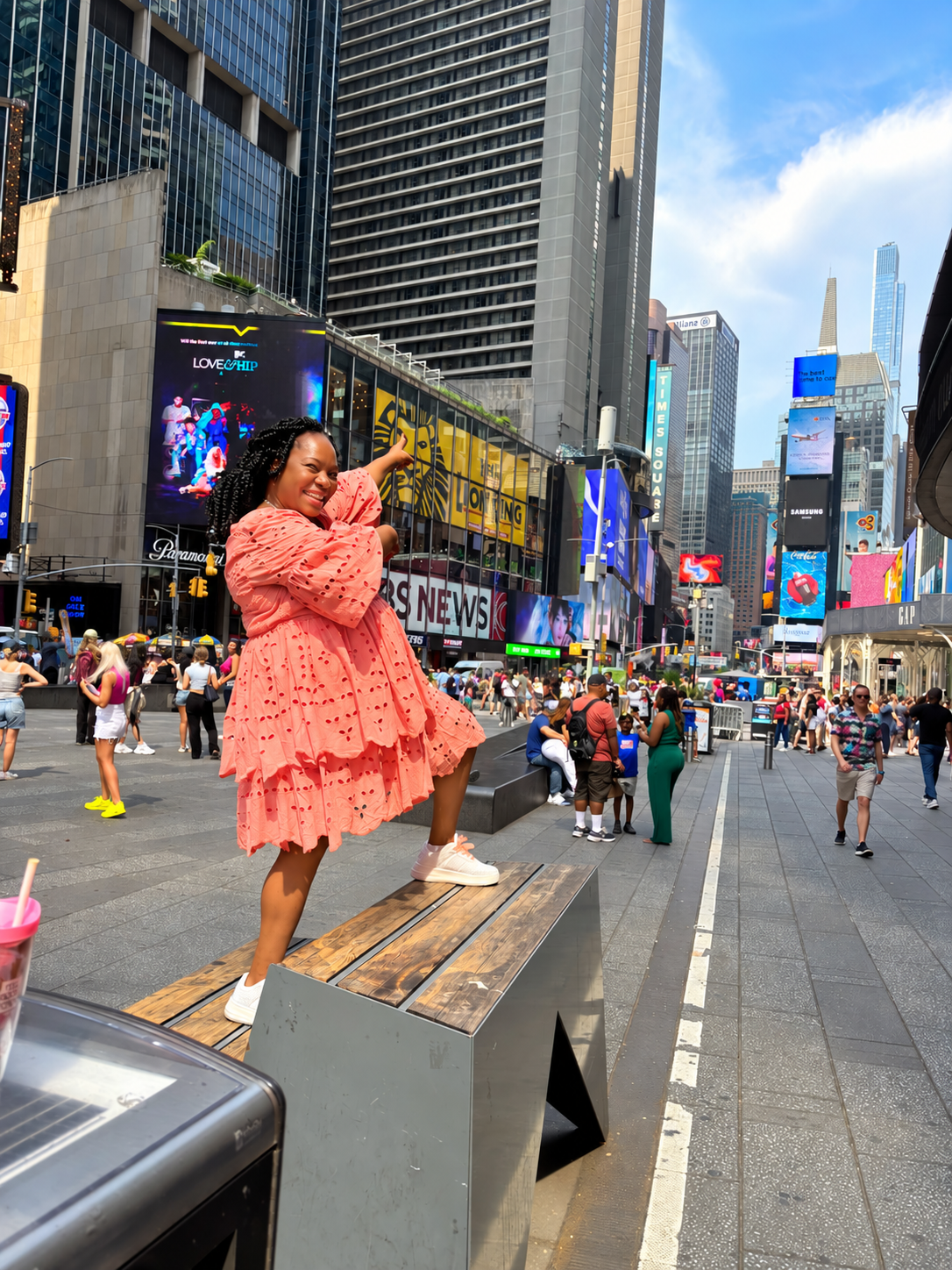 A smiling woman in a pink dress with black hair styled in twists standing on a bench in Times Square, New York City, surrounded by crowds, tall buildings, digital billboards, and neon signs.