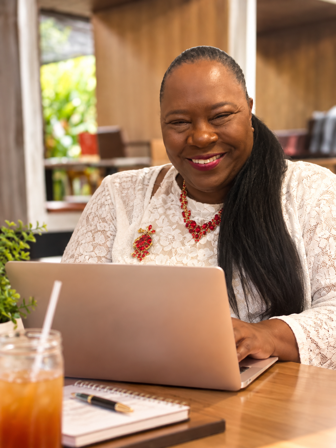 A woman with dark hair, wearing a white lace top and red jewelry, smiling while working on a laptop at a wooden table in a cozy, well-lit café or restaurant.