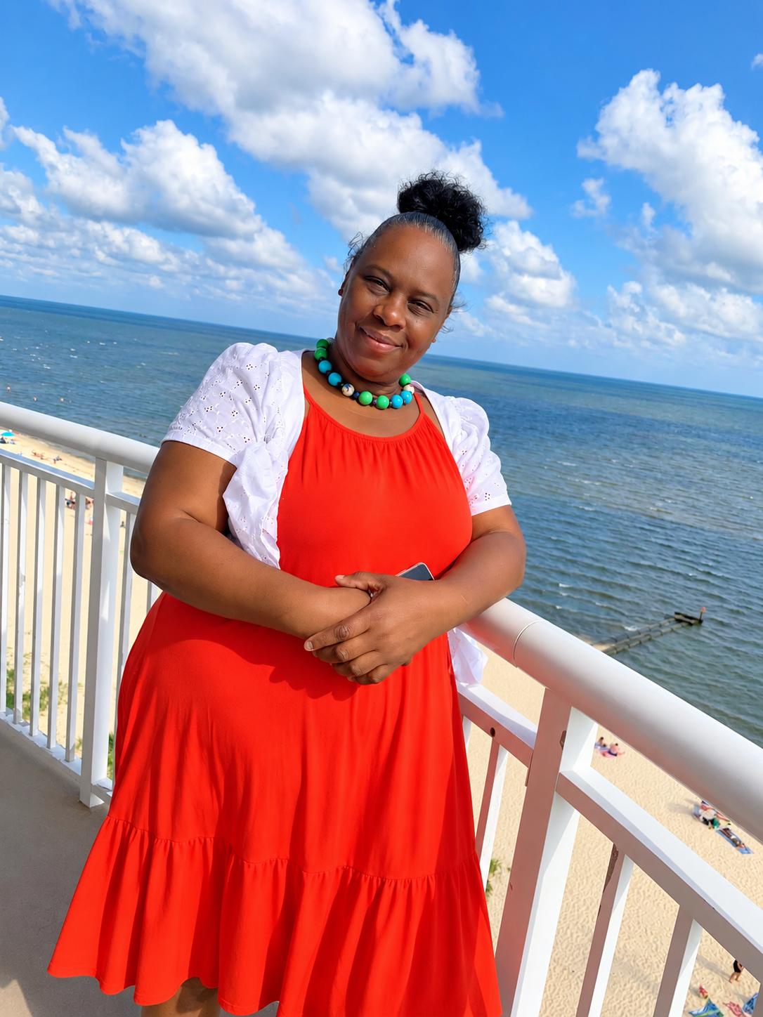 A woman in a red dress and white jacket with a blue and green necklace standing on a balcony by the ocean.