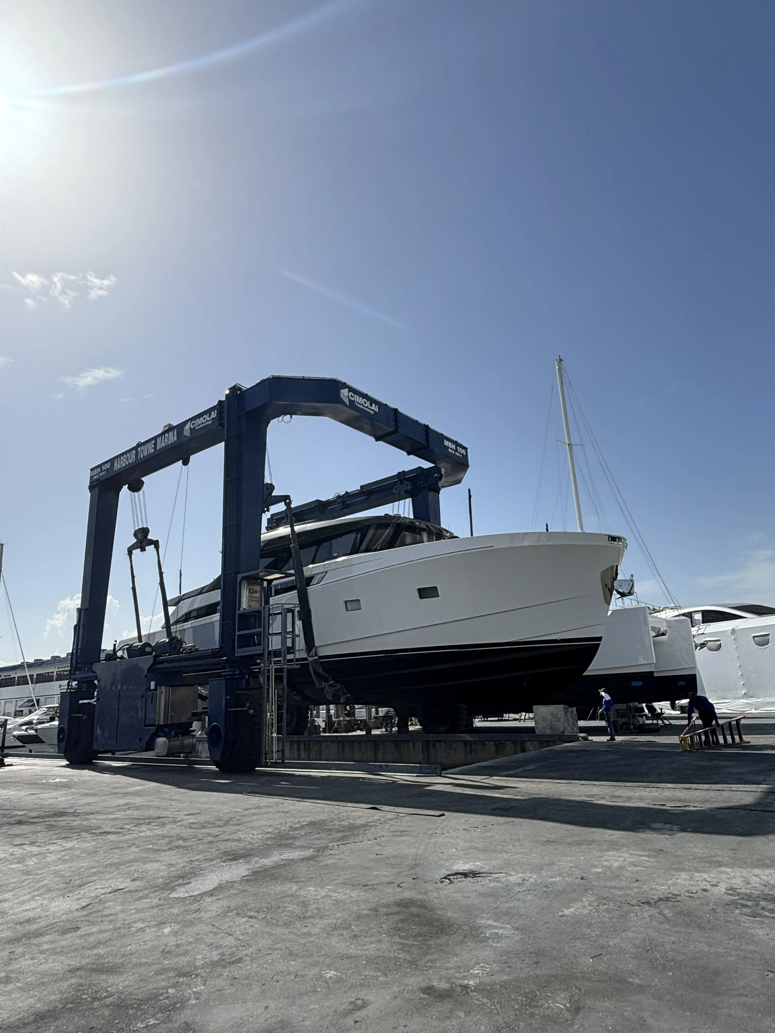 A large boat is being lifted out of the water at a marina using a blue boat lift. The sky is clear with some clouds and sunlight.