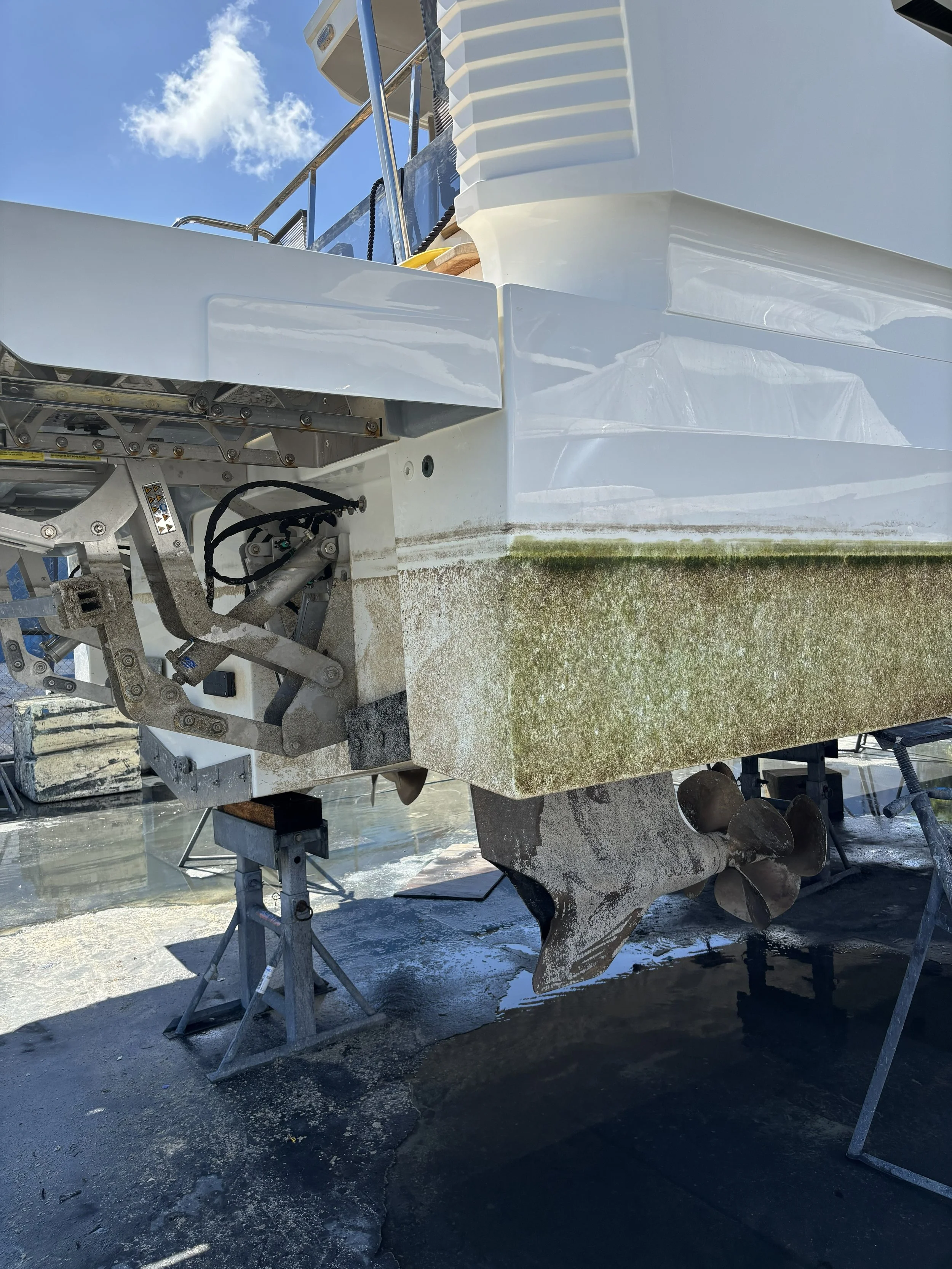 Close-up view of the stern of a white boat on dry land, with a rusted propeller and some algae growth on the hull, supported by metal stands.