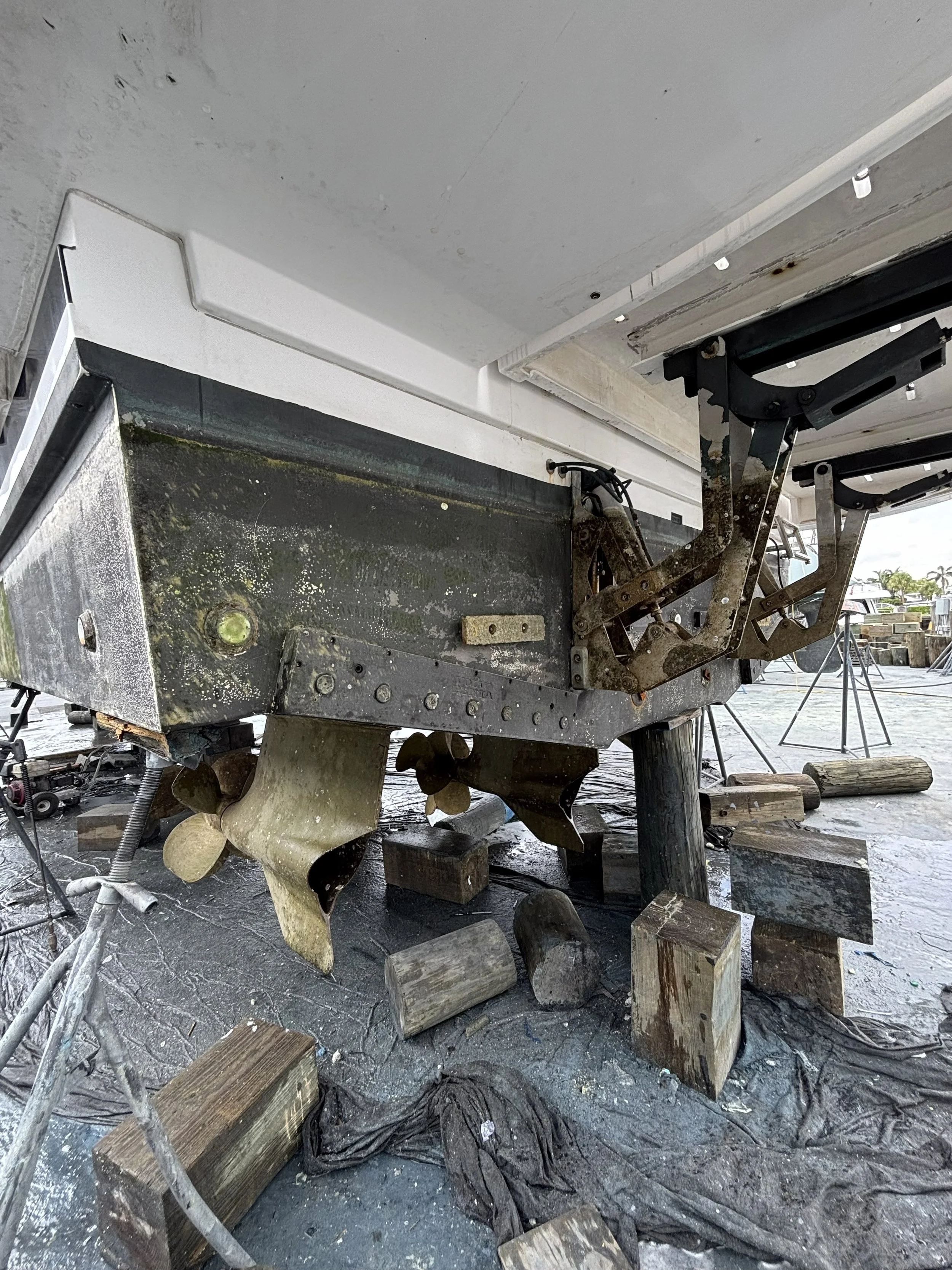 View underneath a boat showing the hull with visible hardware and propeller, surrounded by wooden blocks and scaffolding on a dock.