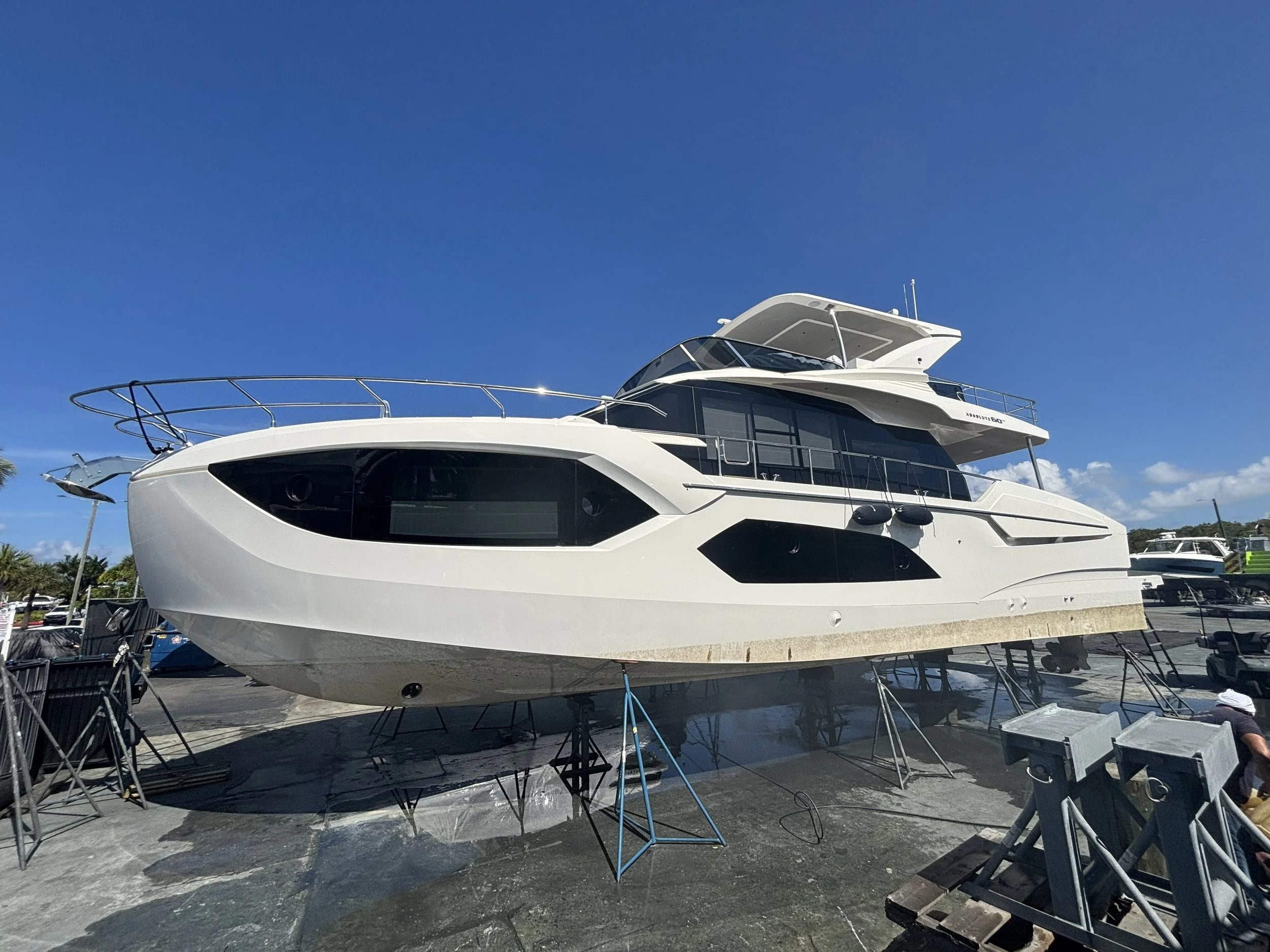 A white luxury yacht is on land, supported by stands, ready for transportation or maintenance. The yacht features multiple decks, large windows, and a sleek modern design. The background includes a clear blue sky, indicating a sunny day.