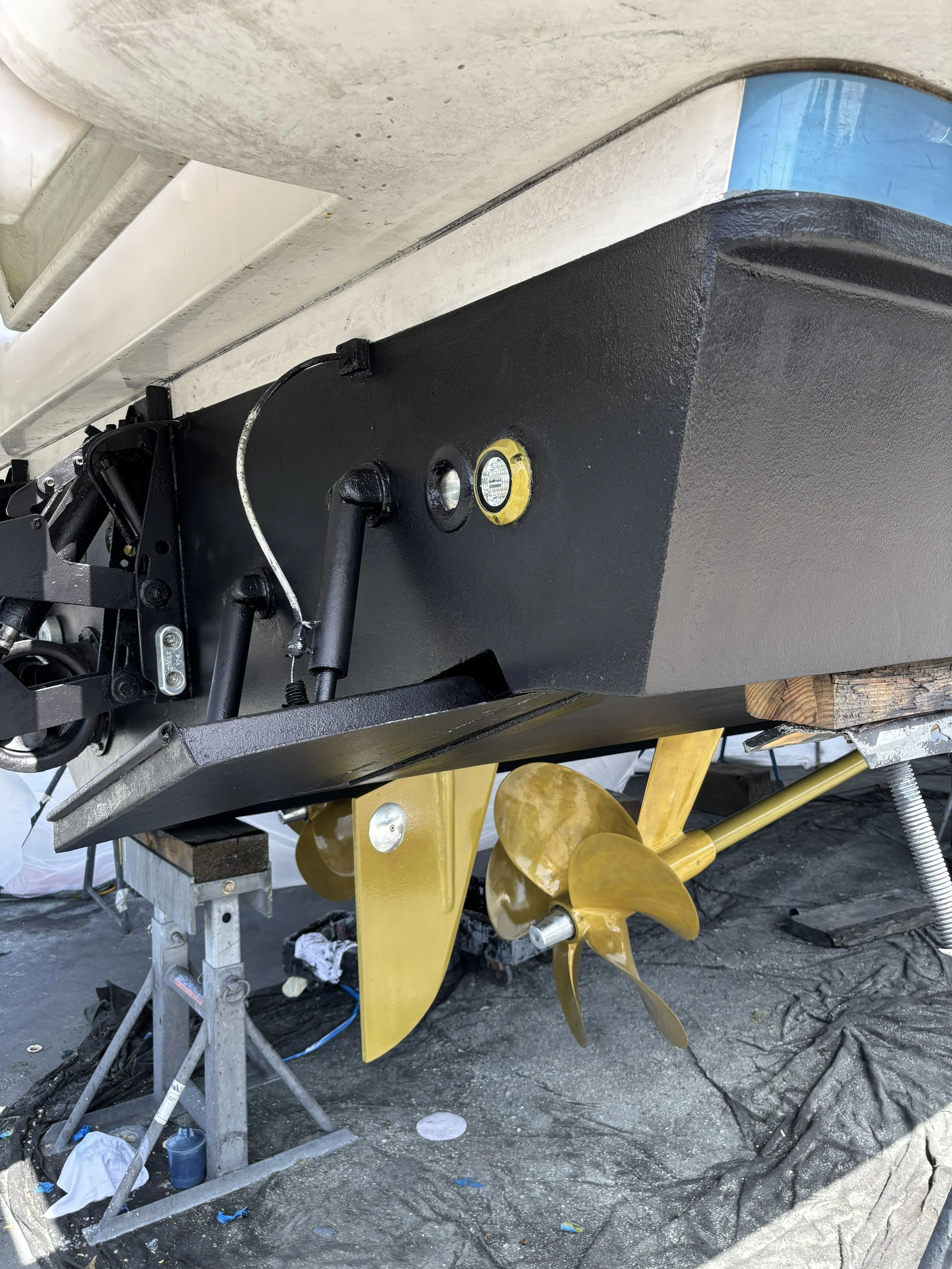 Close-up view of a boat propeller with four yellow blades, mounted beneath a boat hull supported by a metal stand. Various tools and equipment are visible around the area, indicating maintenance work.