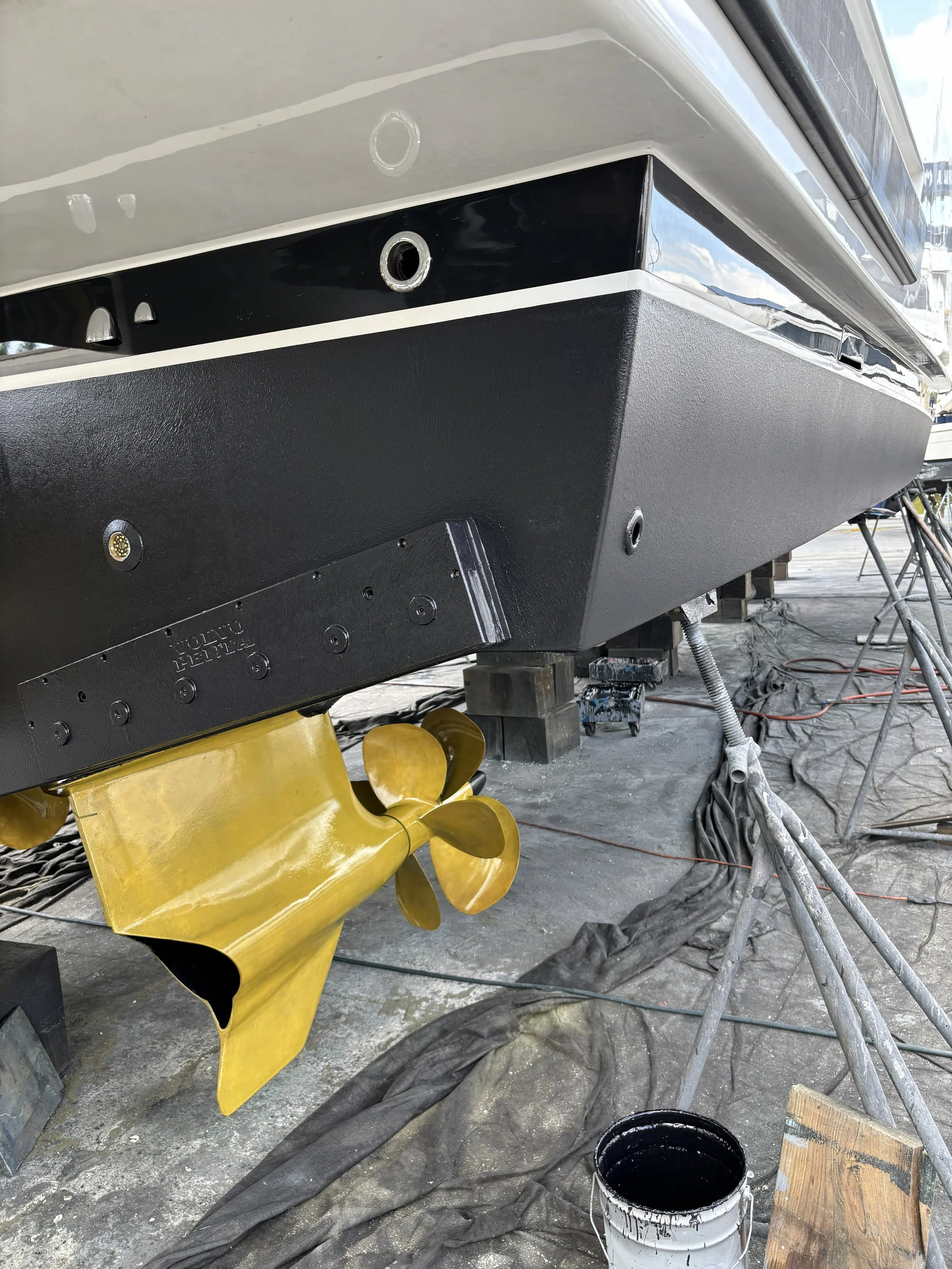 A boat hull under construction with a black bottom and a yellow propeller, surrounded by building tools, black tarp, and a paint bucket.