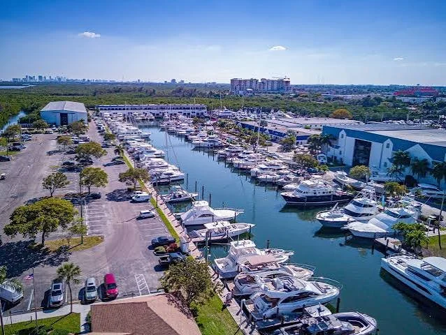 Aerial view of Harbour Towne marina with numerous yachts docked, surrounded by parking lots, trees, buildings, and a distant city skyline under a blue sky.
